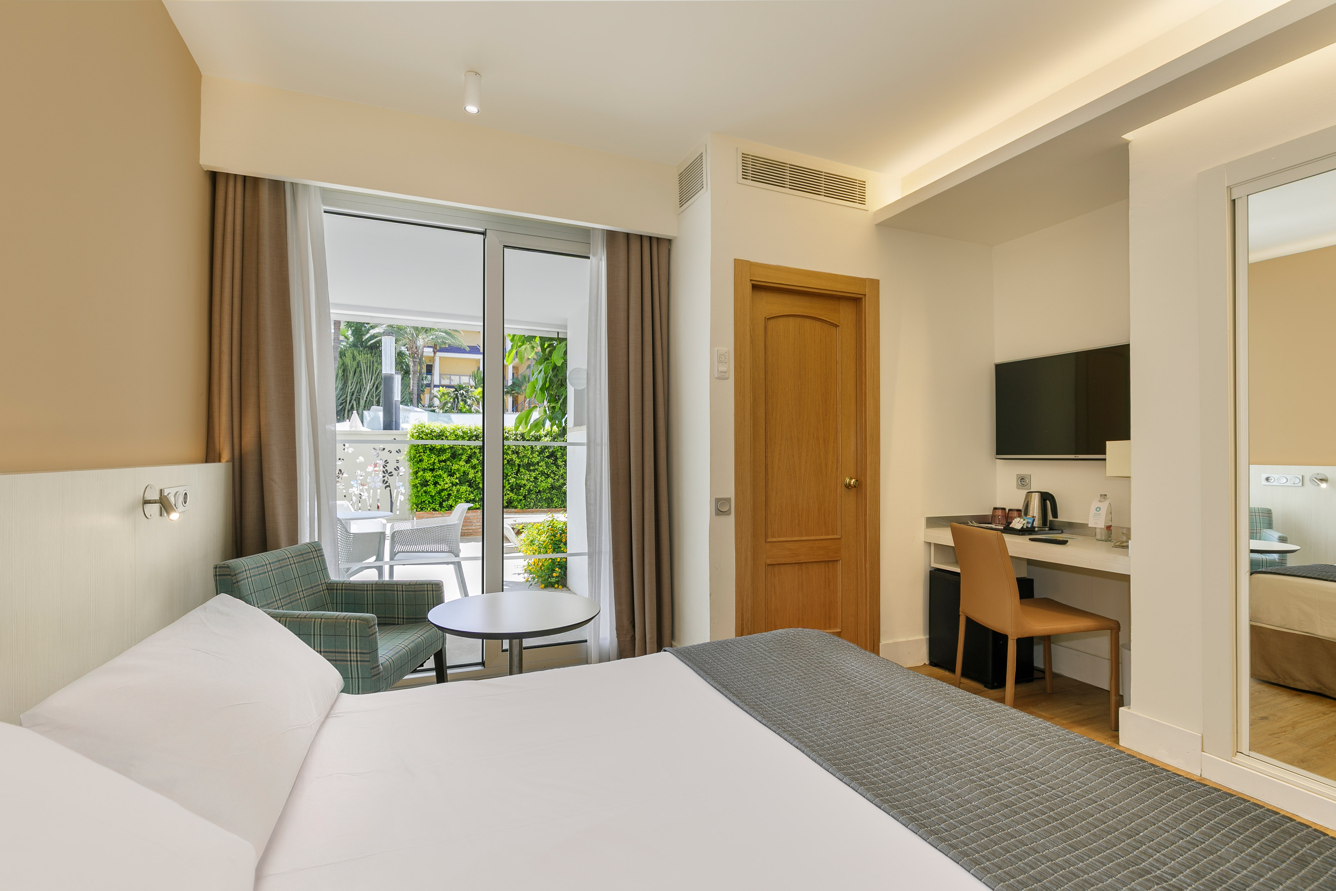 Modern hotel room with bed, desk, balcony, and lush garden view.