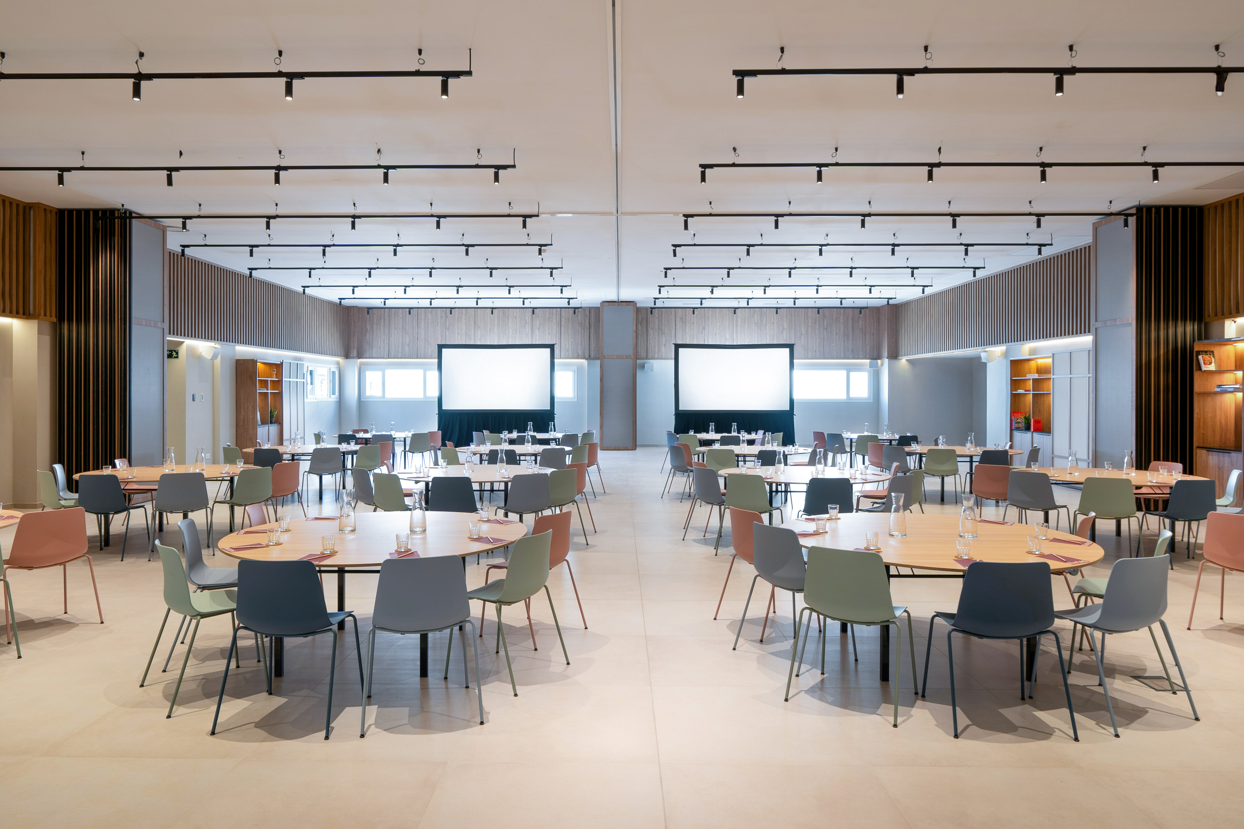 Modern meeting room setup with colorful chairs, round tables, and dual presentation screens.
