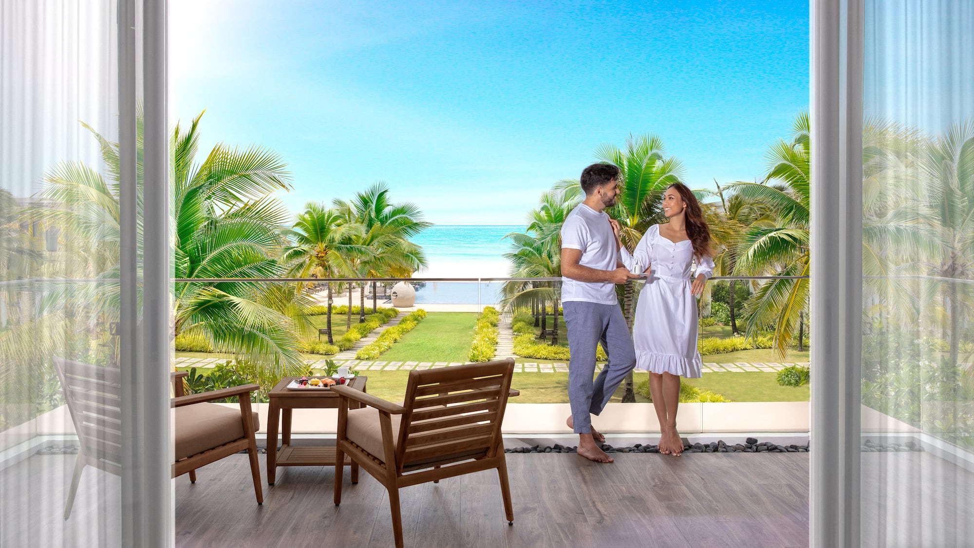 Couple on balcony overlooking tropical beach, ocean, and palm trees.