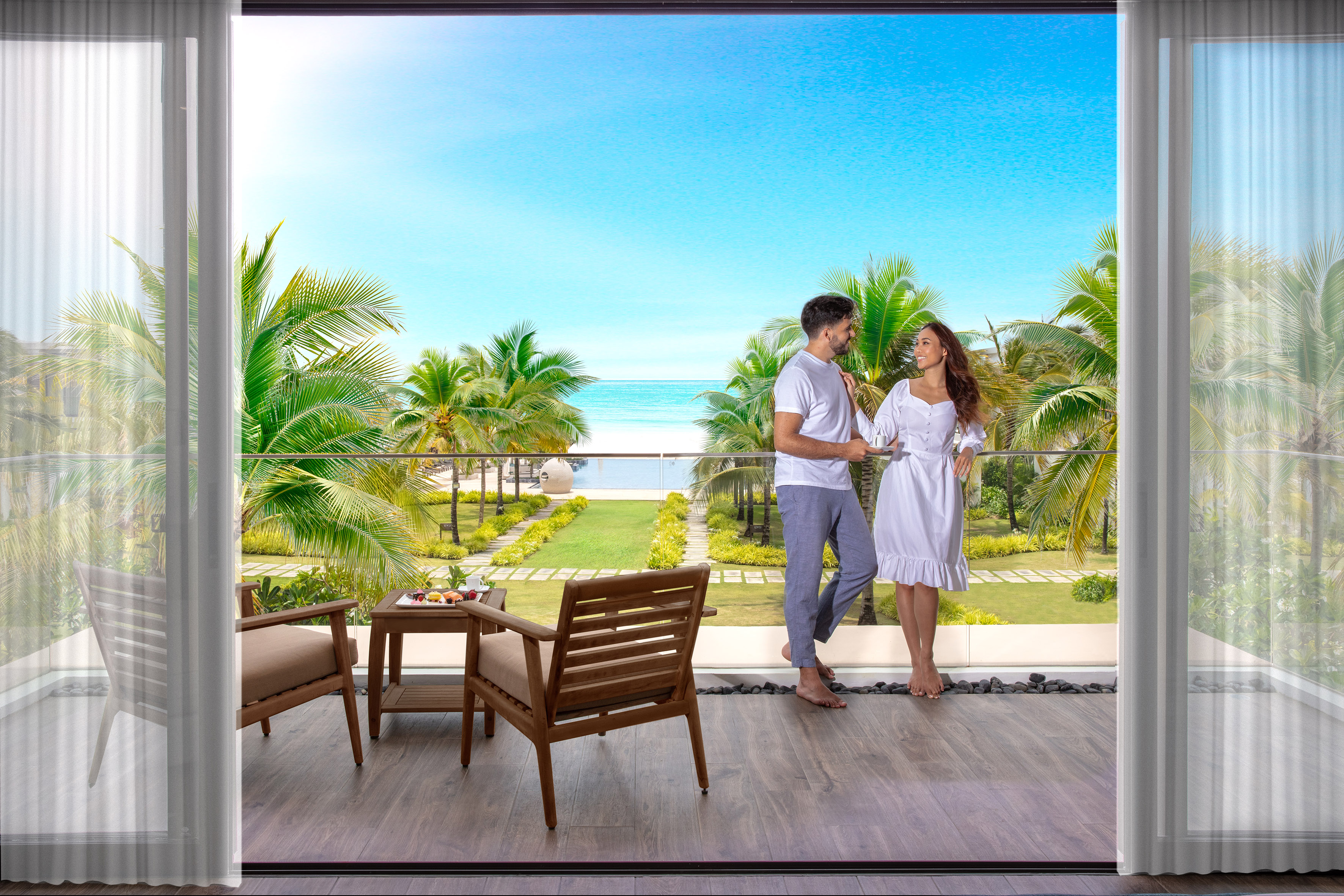 Couple on balcony overlooking tropical beach, ocean, and palm trees.
