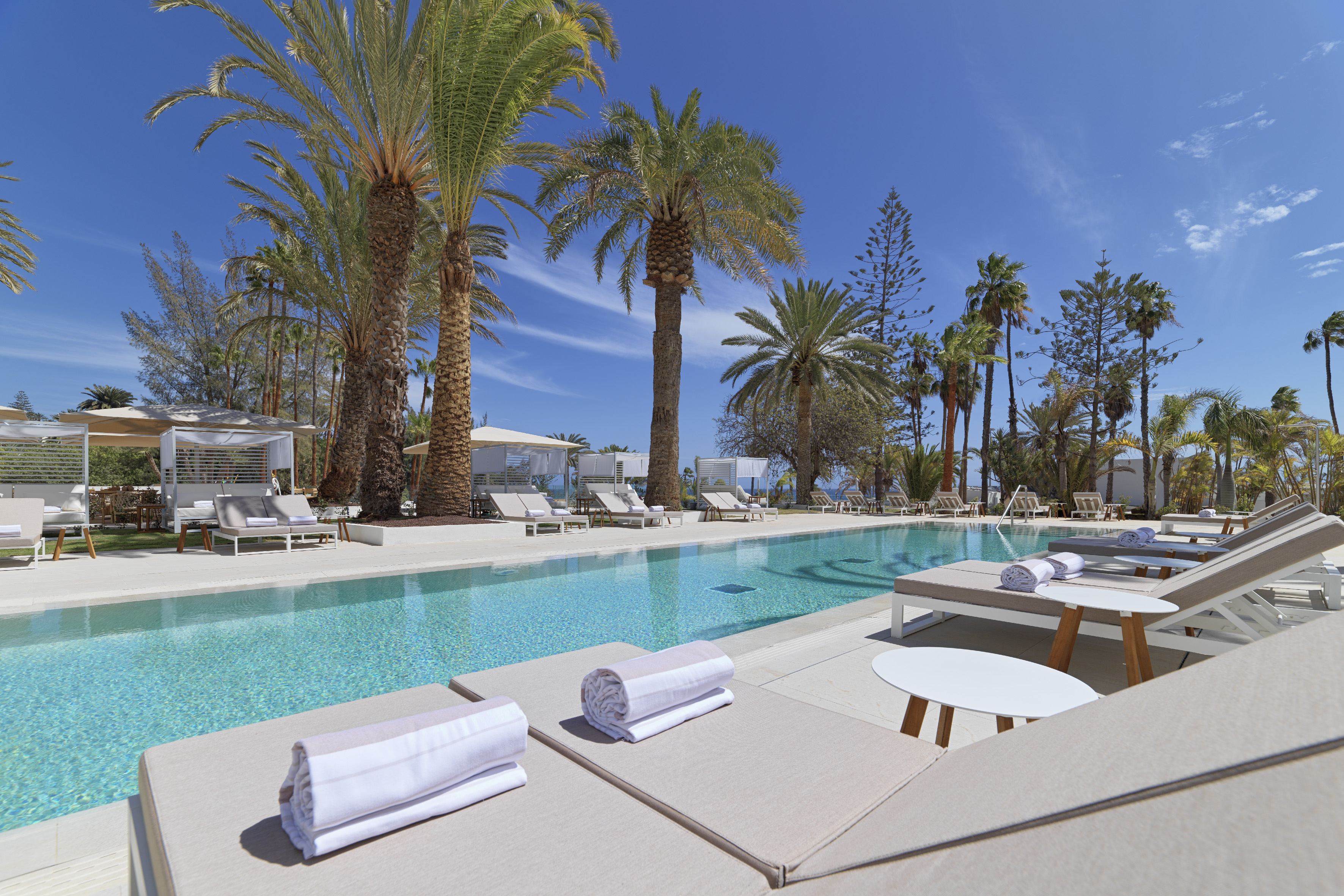 Luxury resort pool, palm trees, sun loungers, white cabanas, and blue sky.