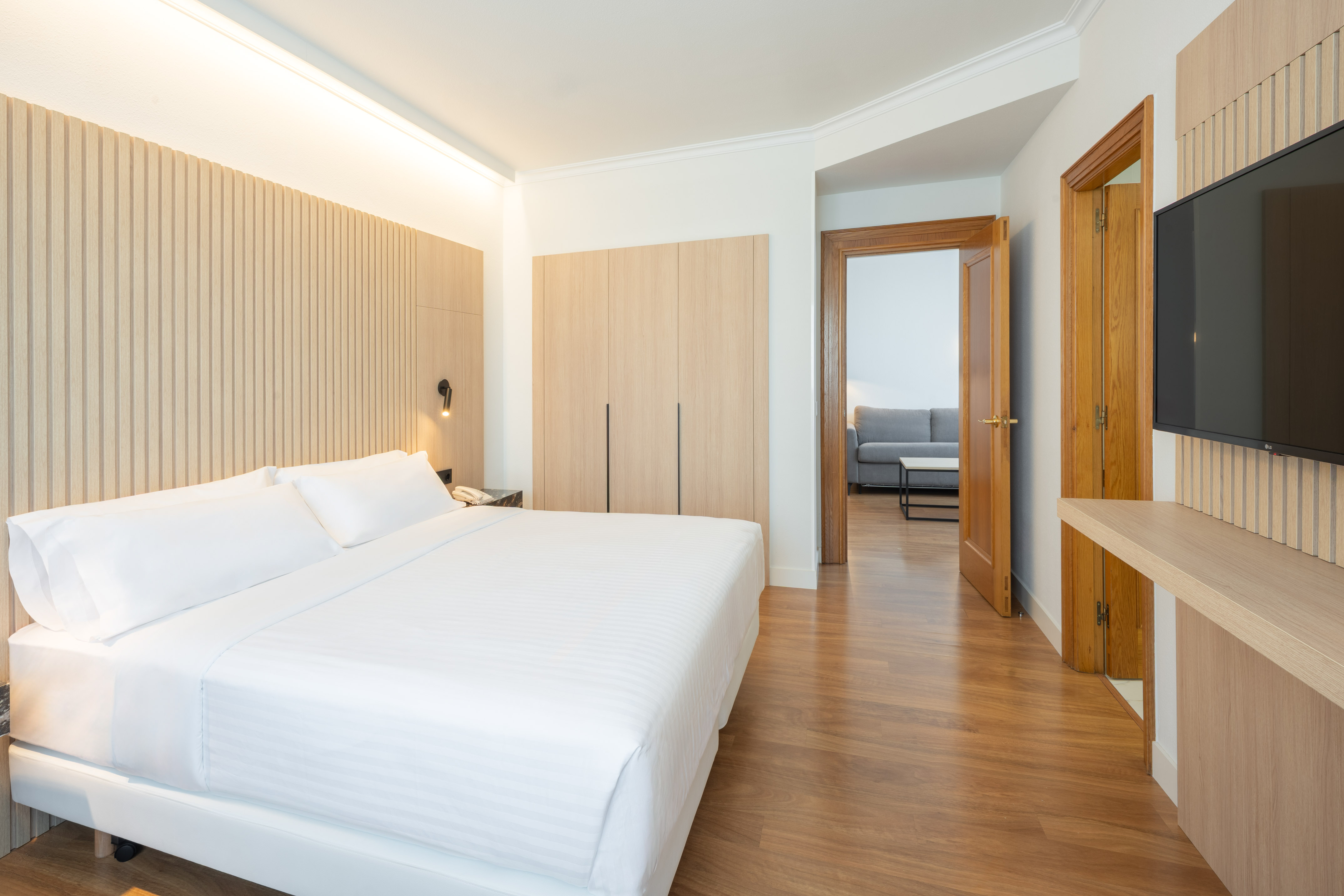 Modern hotel suite bedroom with white bed, wood walls, and hardwood floor.