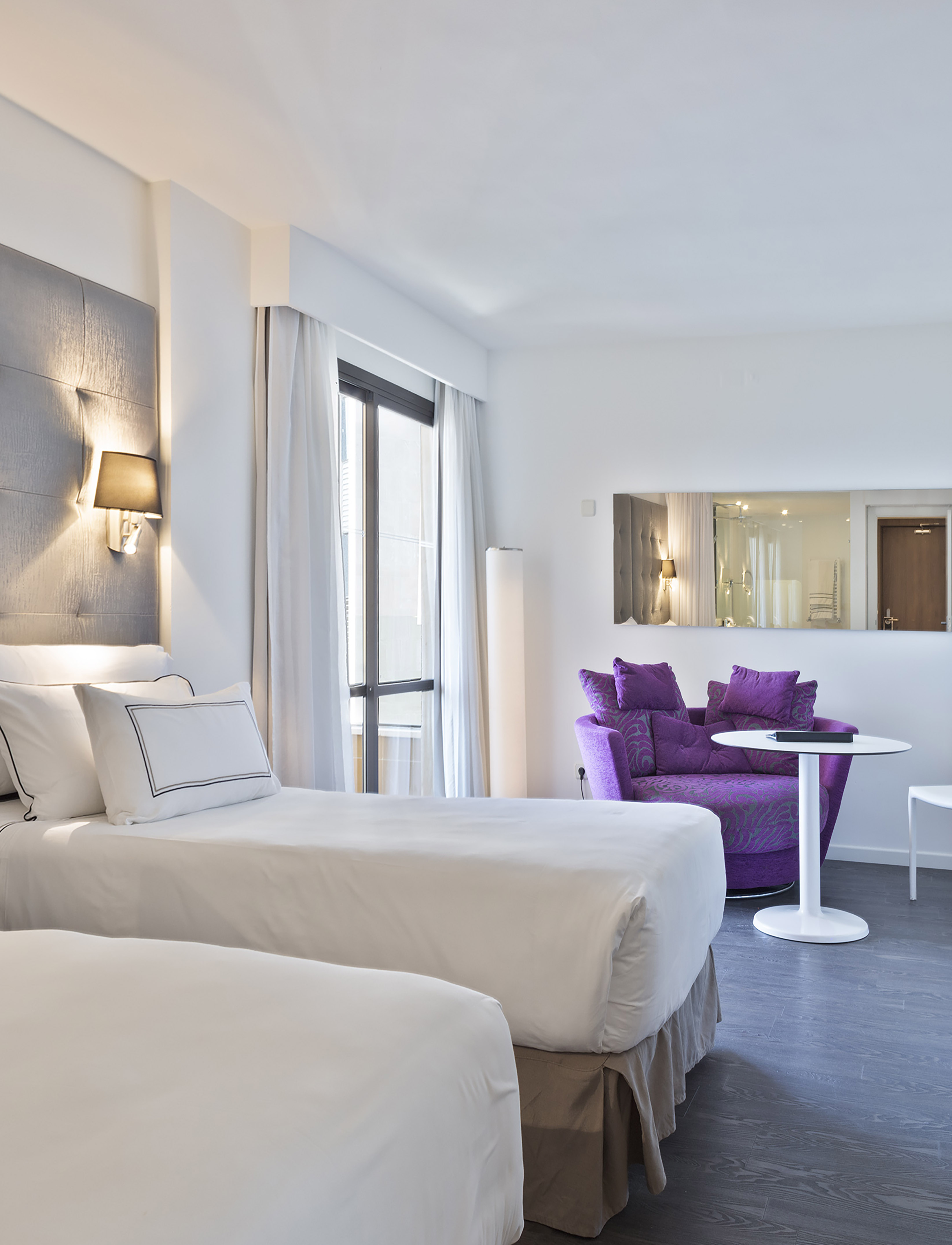 Contemporary hotel room with twin beds, purple accent chair, and large window.