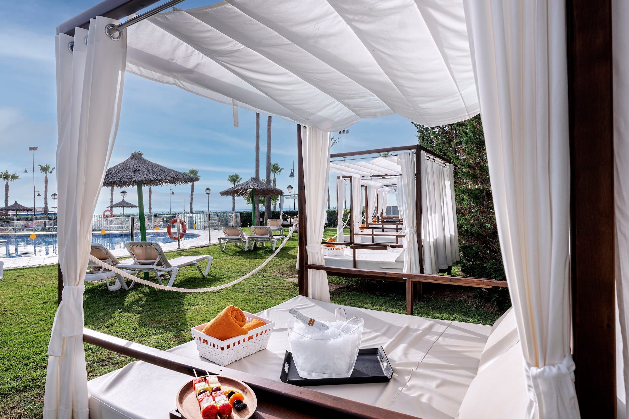 Luxury resort cabana with champagne, fruit, pool, and palm trees.