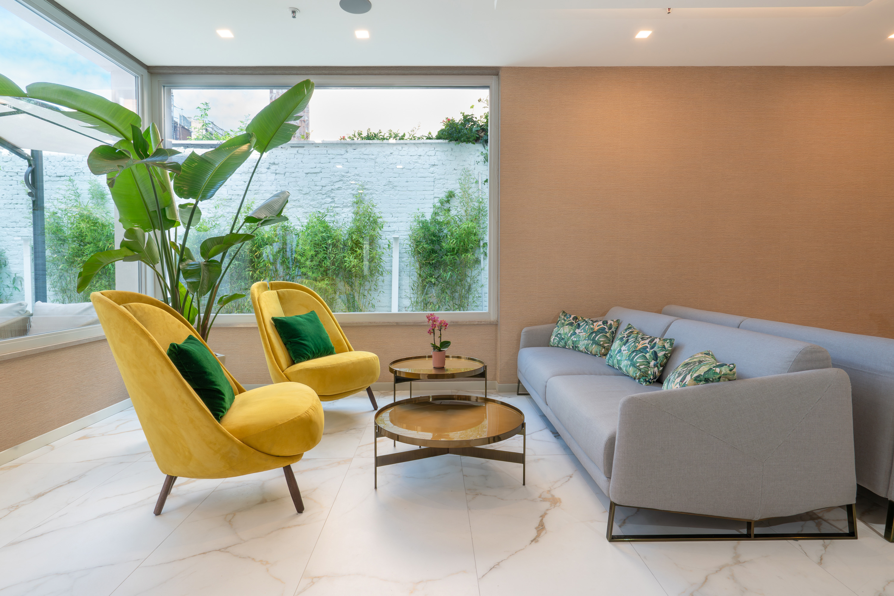 Modern lounge with yellow armchairs, grey sofa, marble floor, and tropical plants.