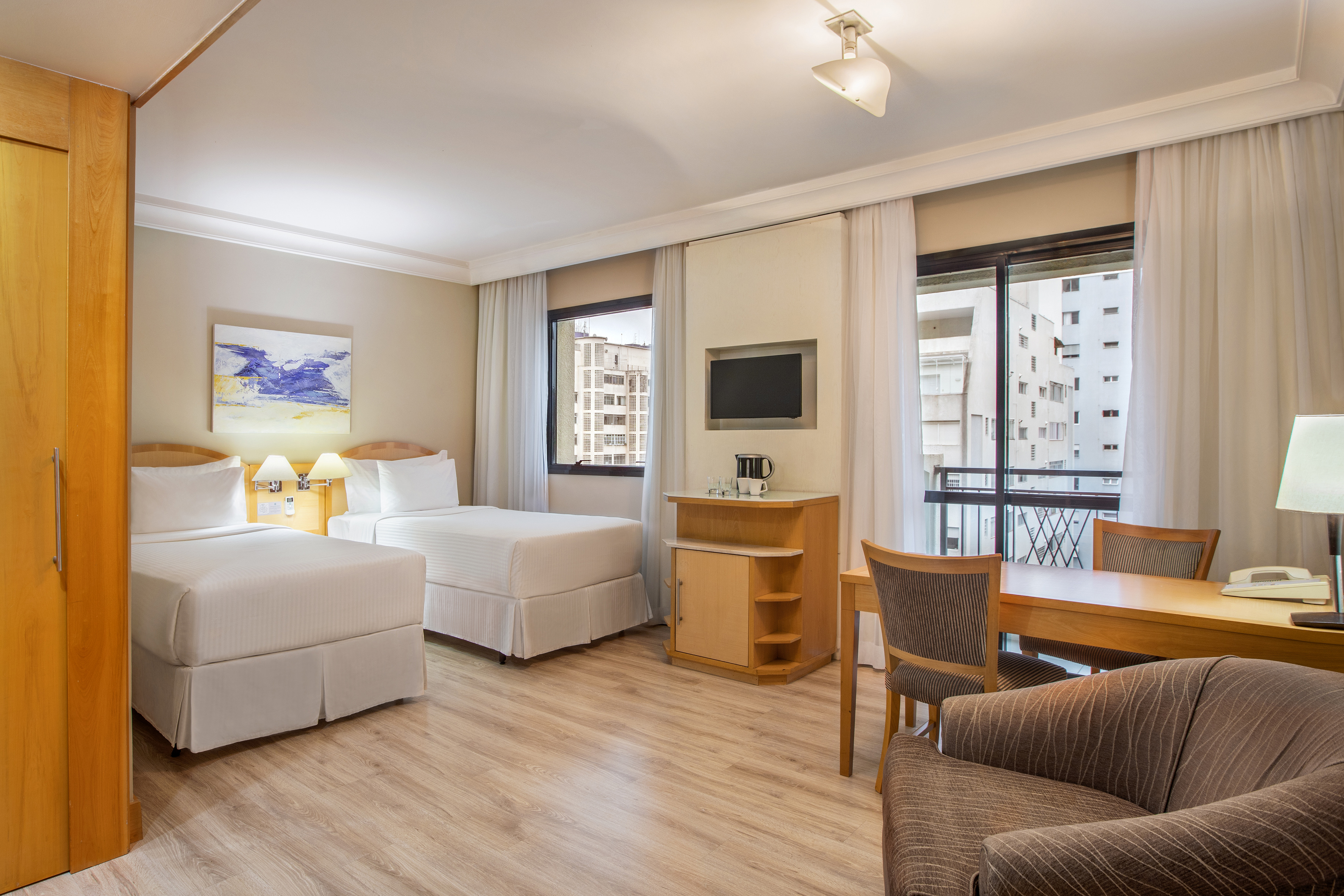 Modern guest room with twin beds, wooden desk, TV, and city view window.