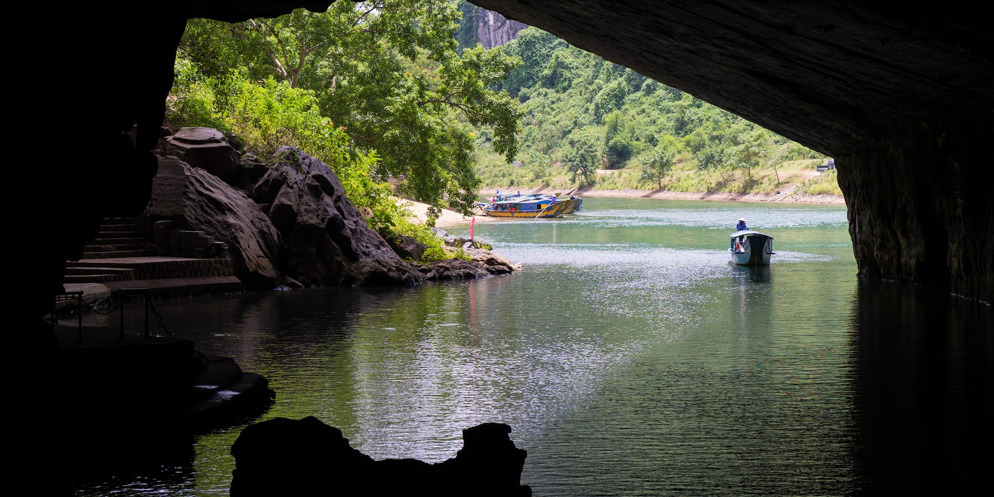 River cave entrance with boats on green water and lush mountains.