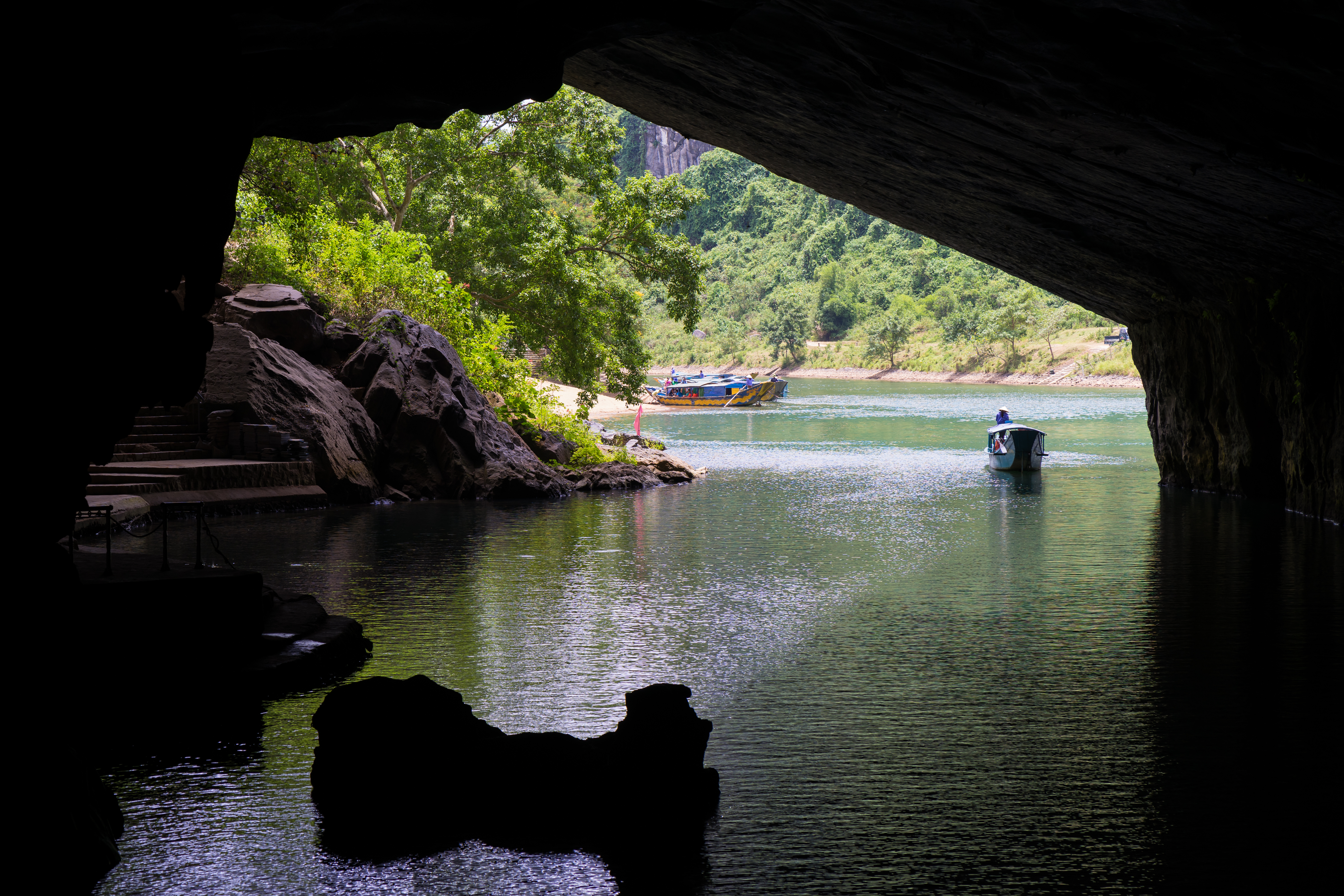 River cave entrance with boats on green water and lush mountains.