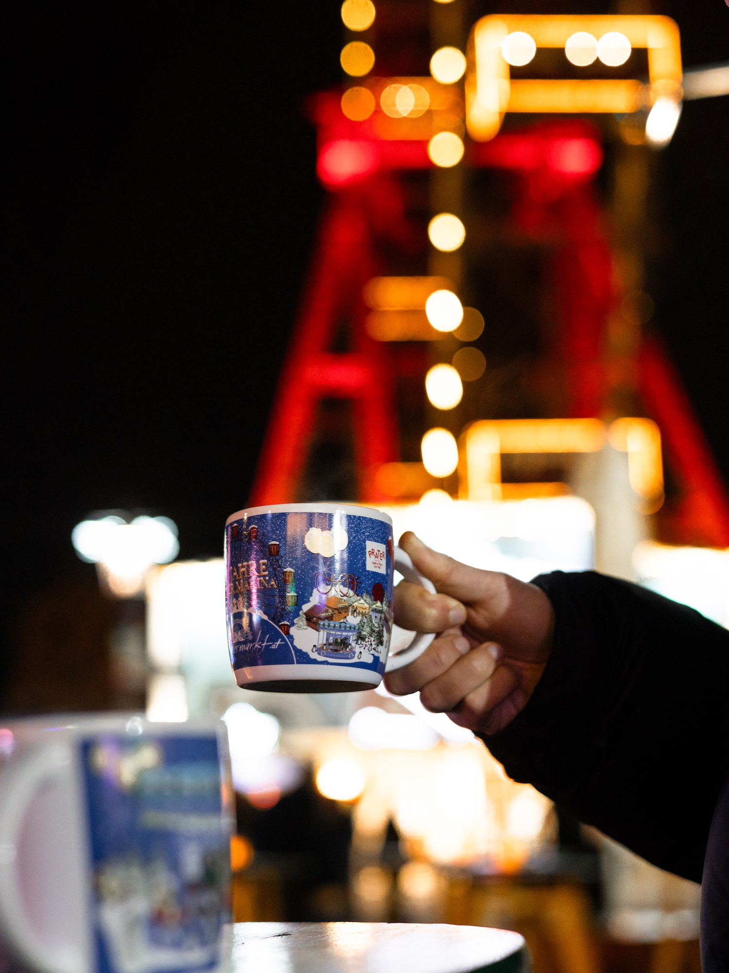 Holding a festive market mug, blurred Ferris wheel lights at night.