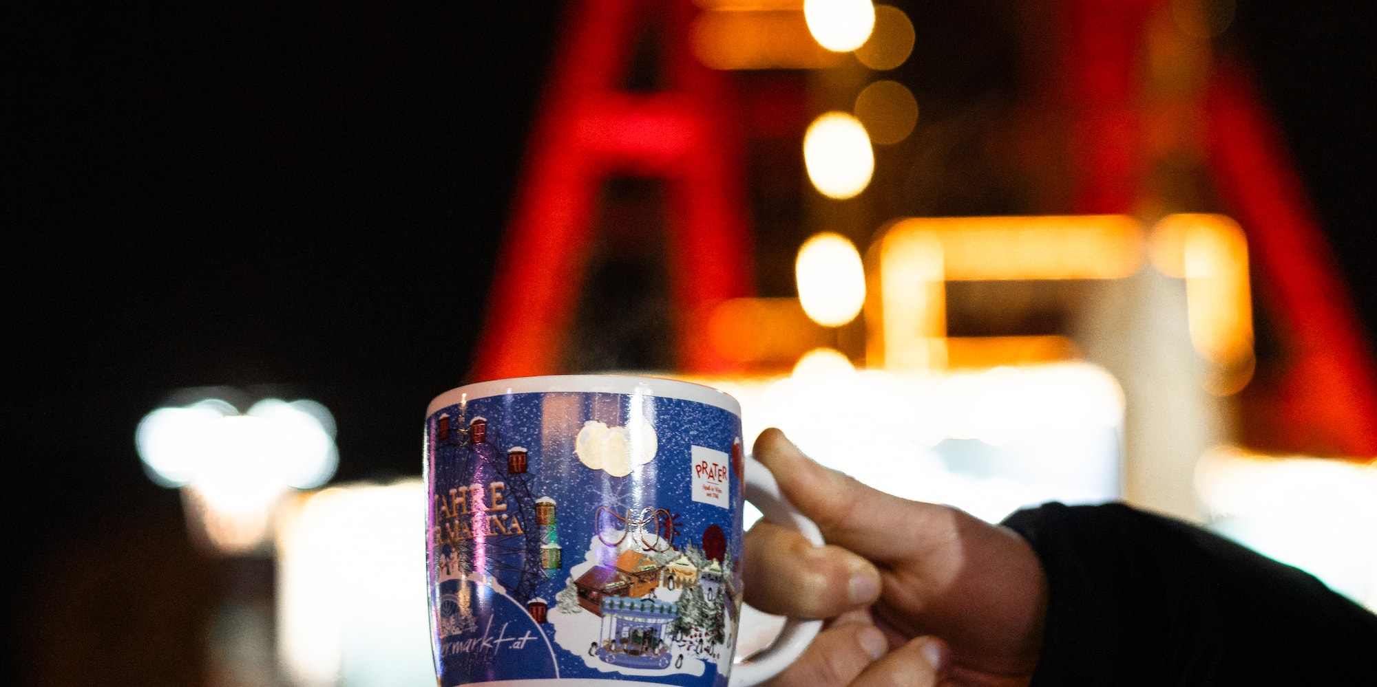 Hand holding blue Christmas market mug with Ferris wheel design, festive lights.