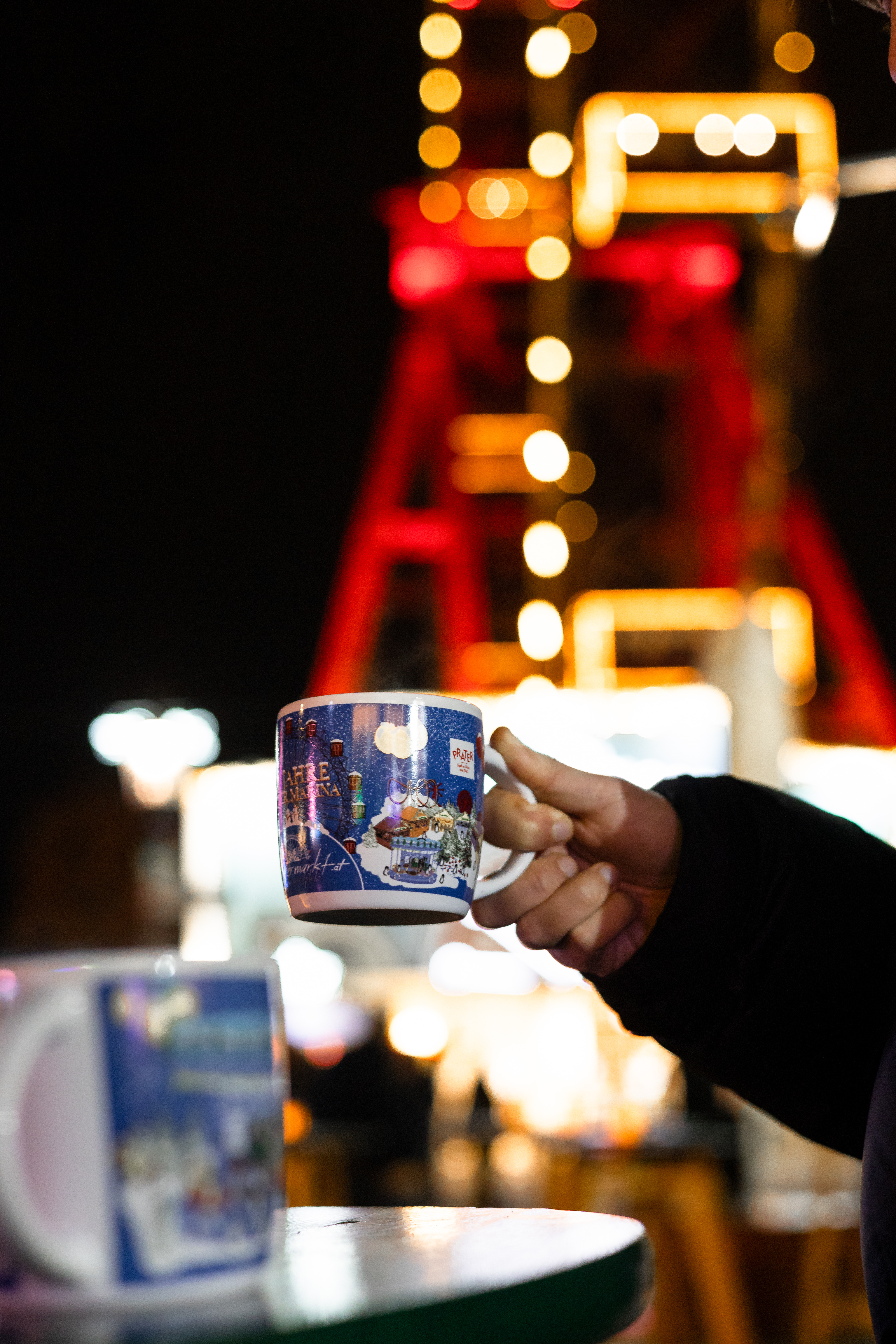 Hand holding blue Christmas market mug with Ferris wheel design, festive lights.