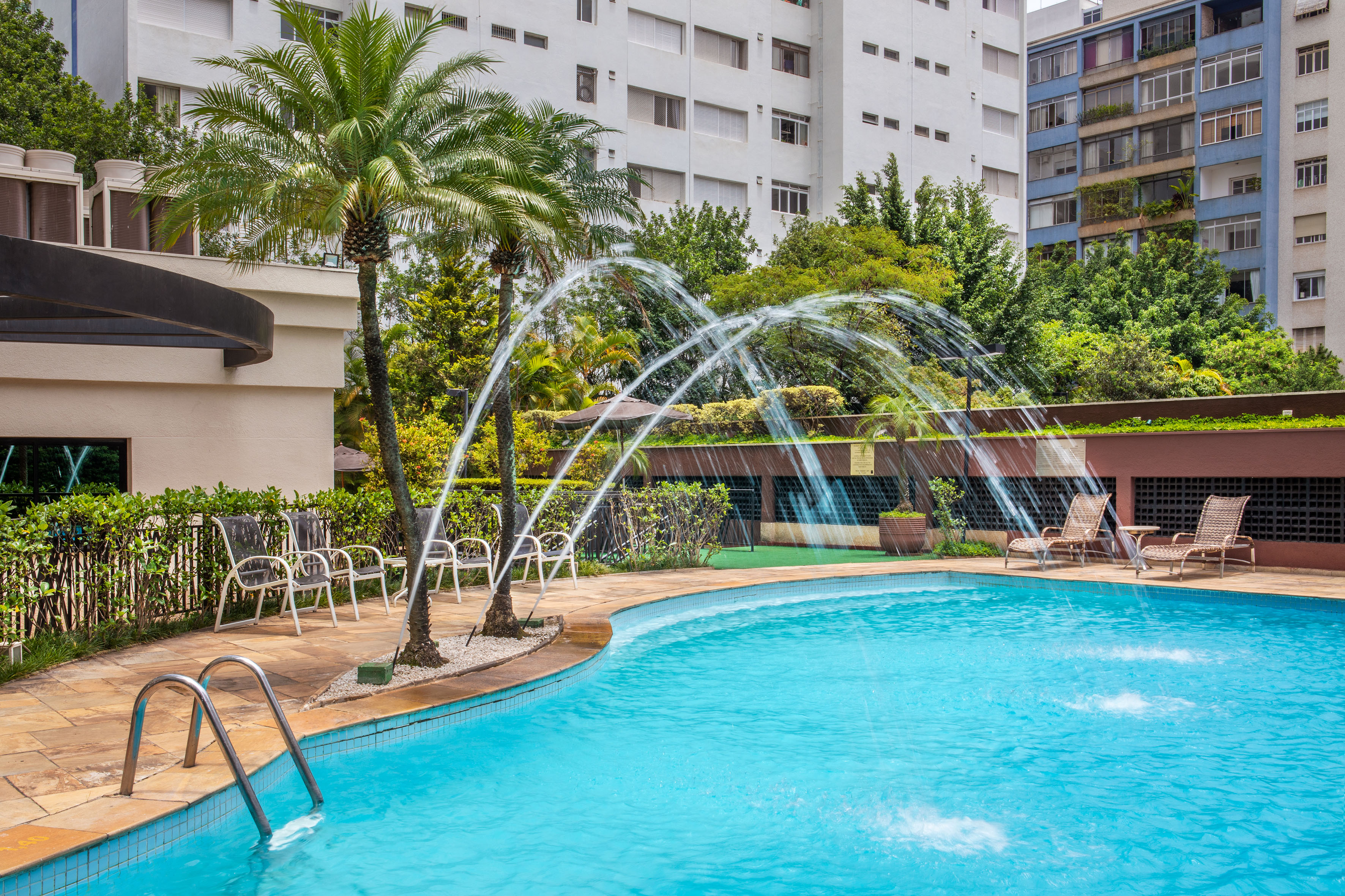 Outdoor swimming pool with water jets, palm trees, and surrounding buildings.