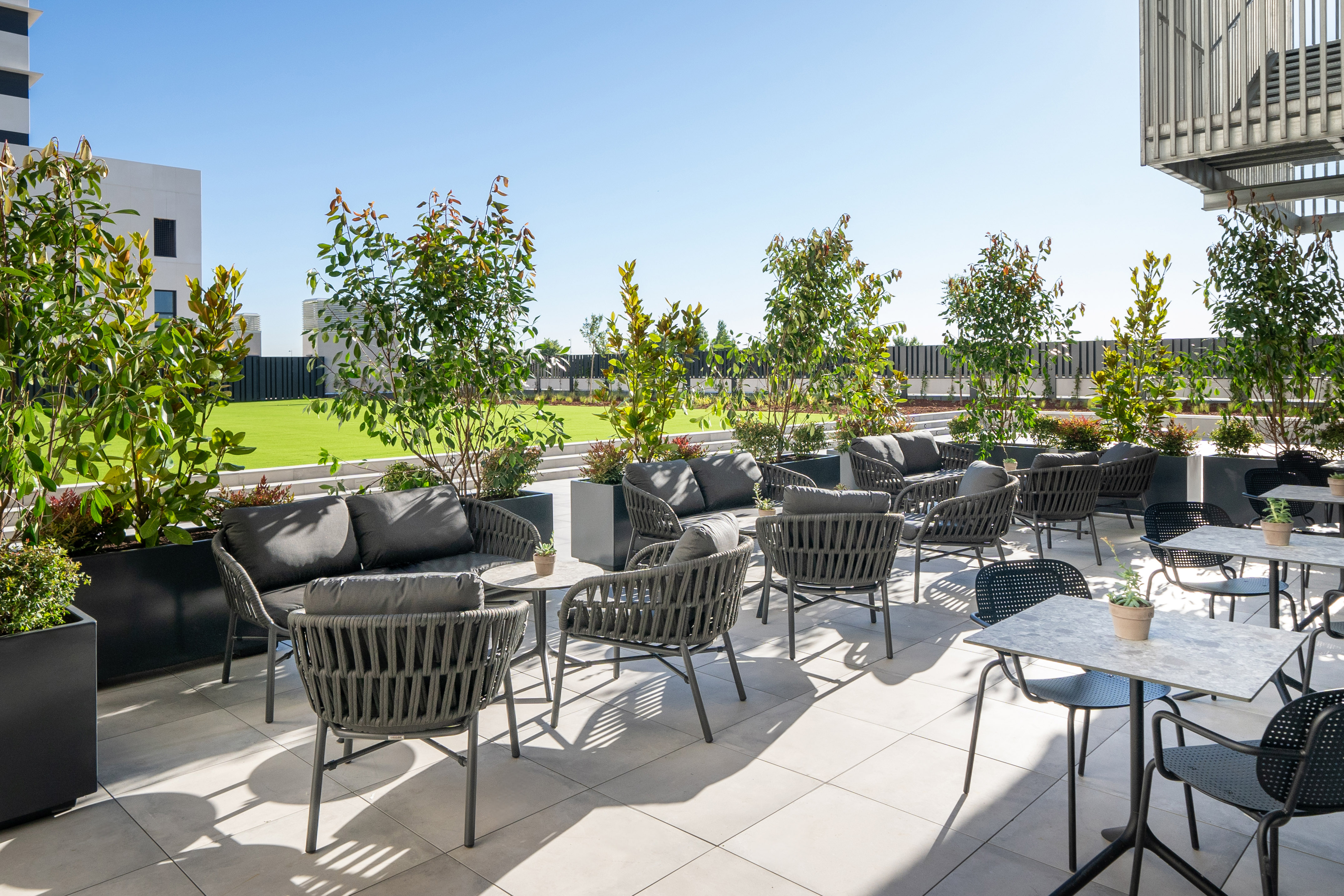 Modern outdoor terrace with grey lounge furniture, tables, plants, and green lawn.