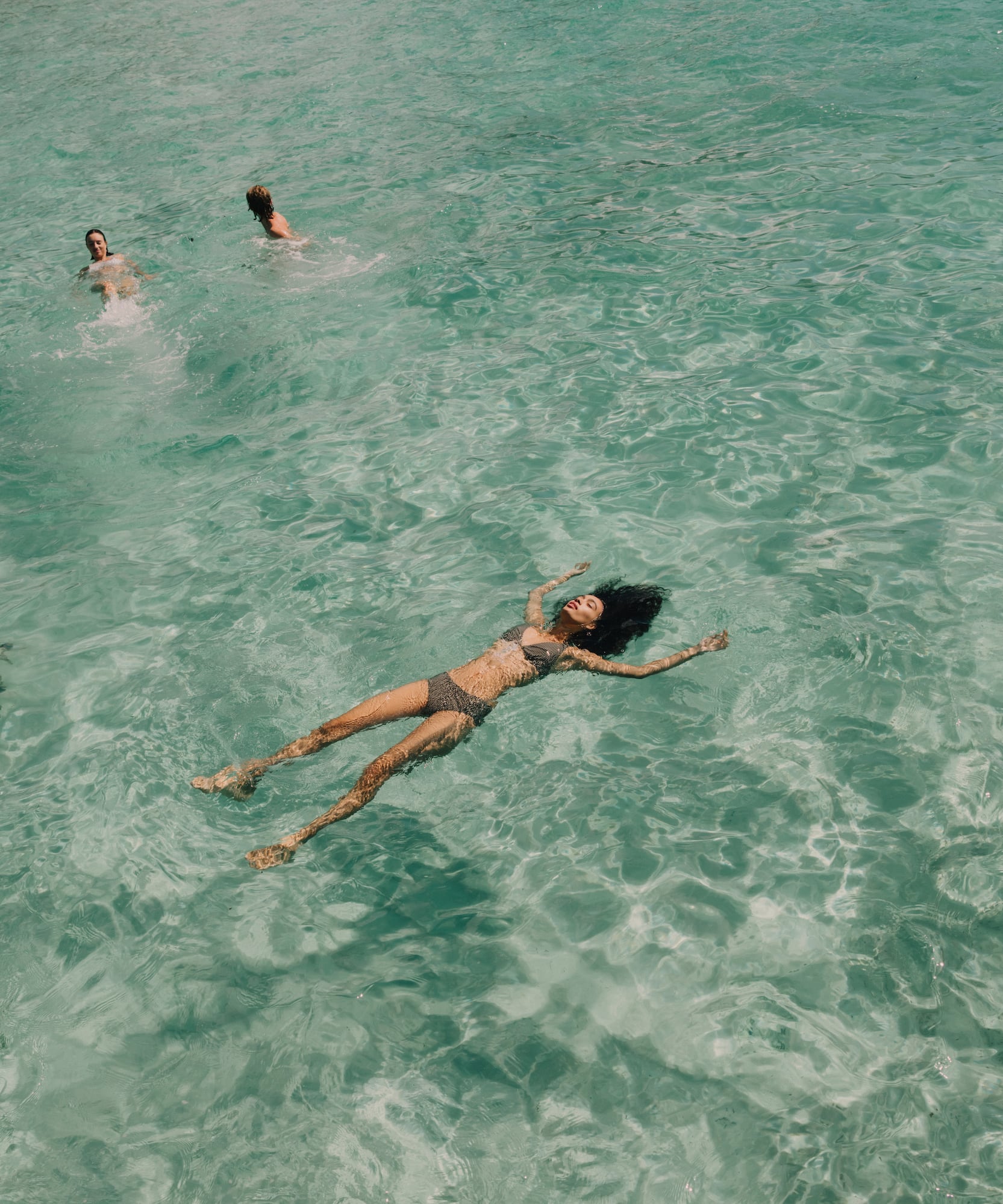 Woman in bikini floating on back in clear turquoise ocean with swimmers.