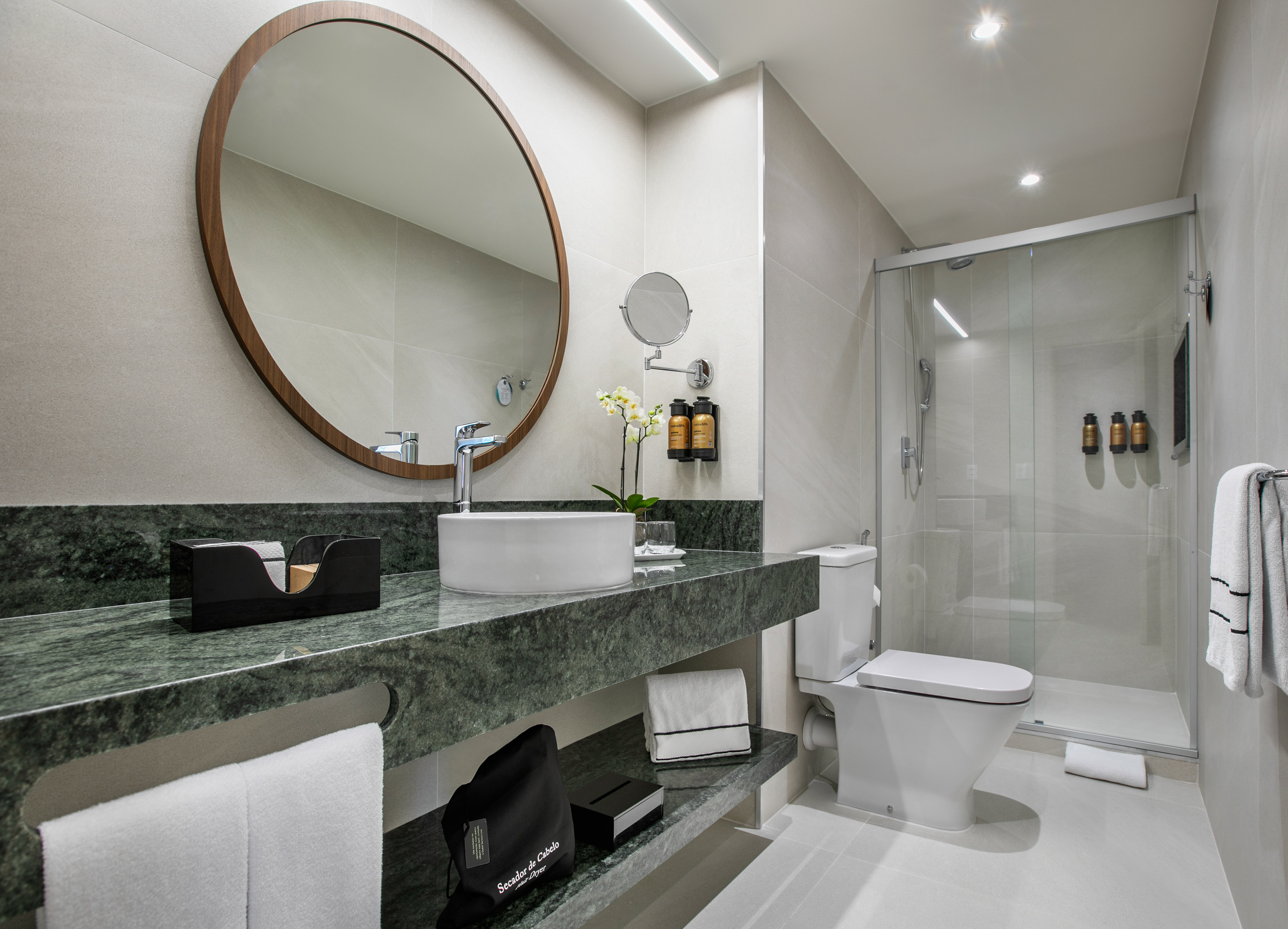 Modern hotel bathroom with green marble vanity, vessel sink, round mirror, and glass shower.
