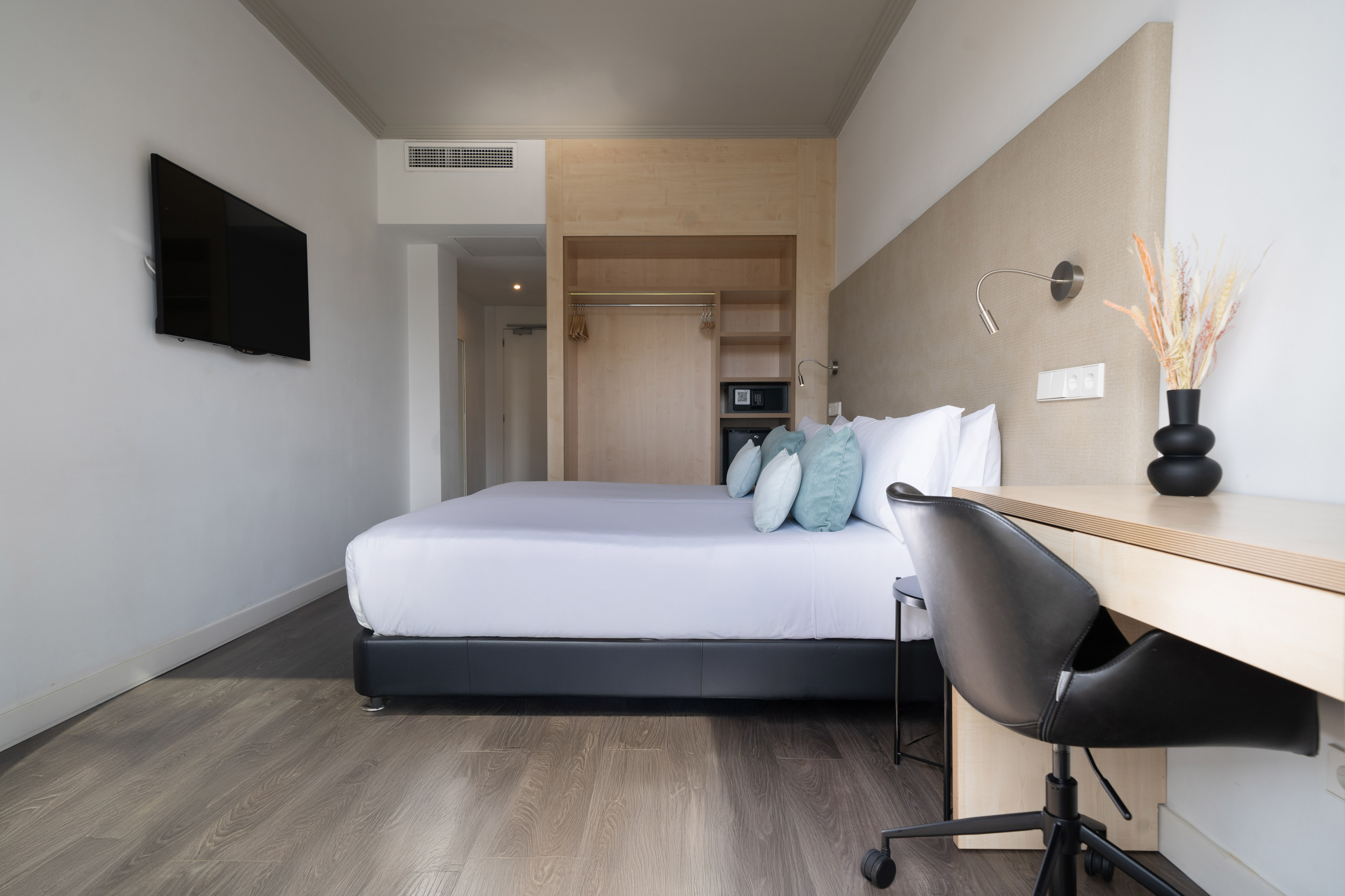 Modern hotel room with bed, desk, wall TV, and integrated wardrobe with safe.