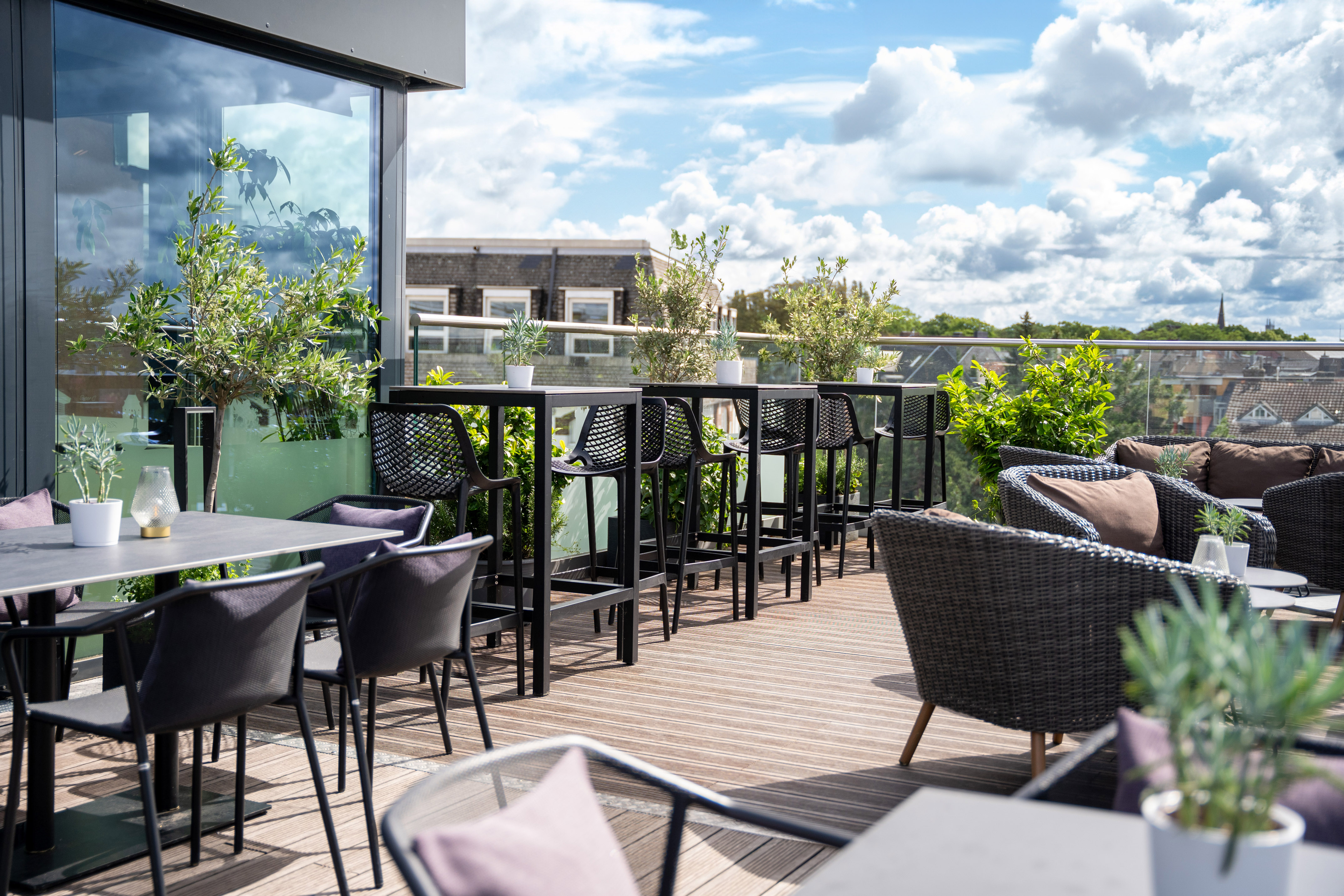 Rooftop terrace with outdoor lounge, bar tables, green plants, and city view.