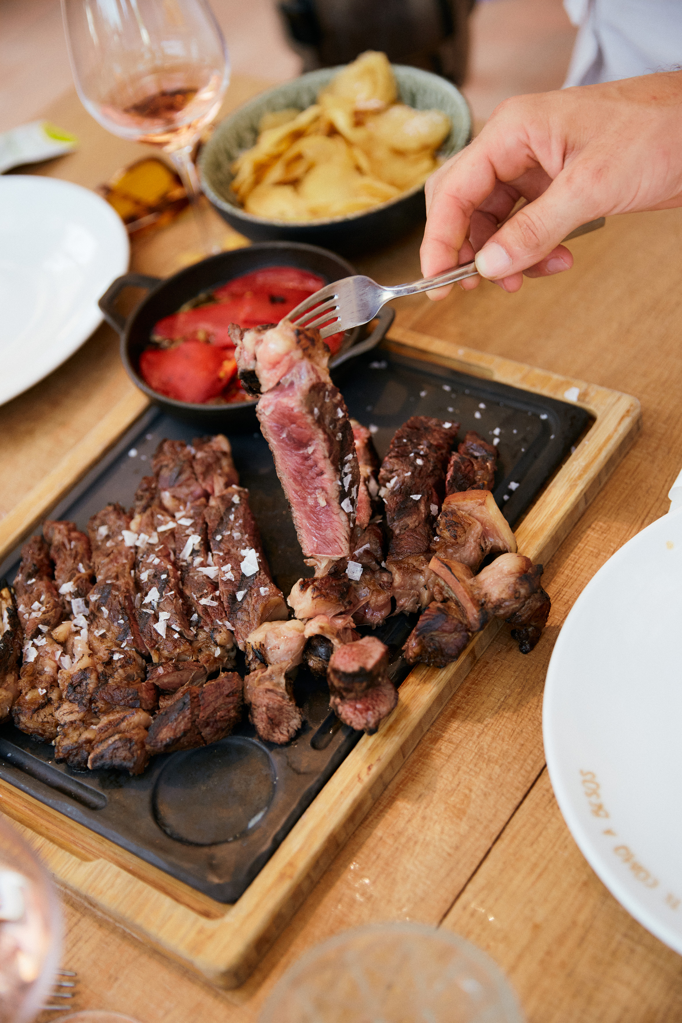 Hand lifting sliced medium-rare steak with sea salt from hot plate, side dishes.