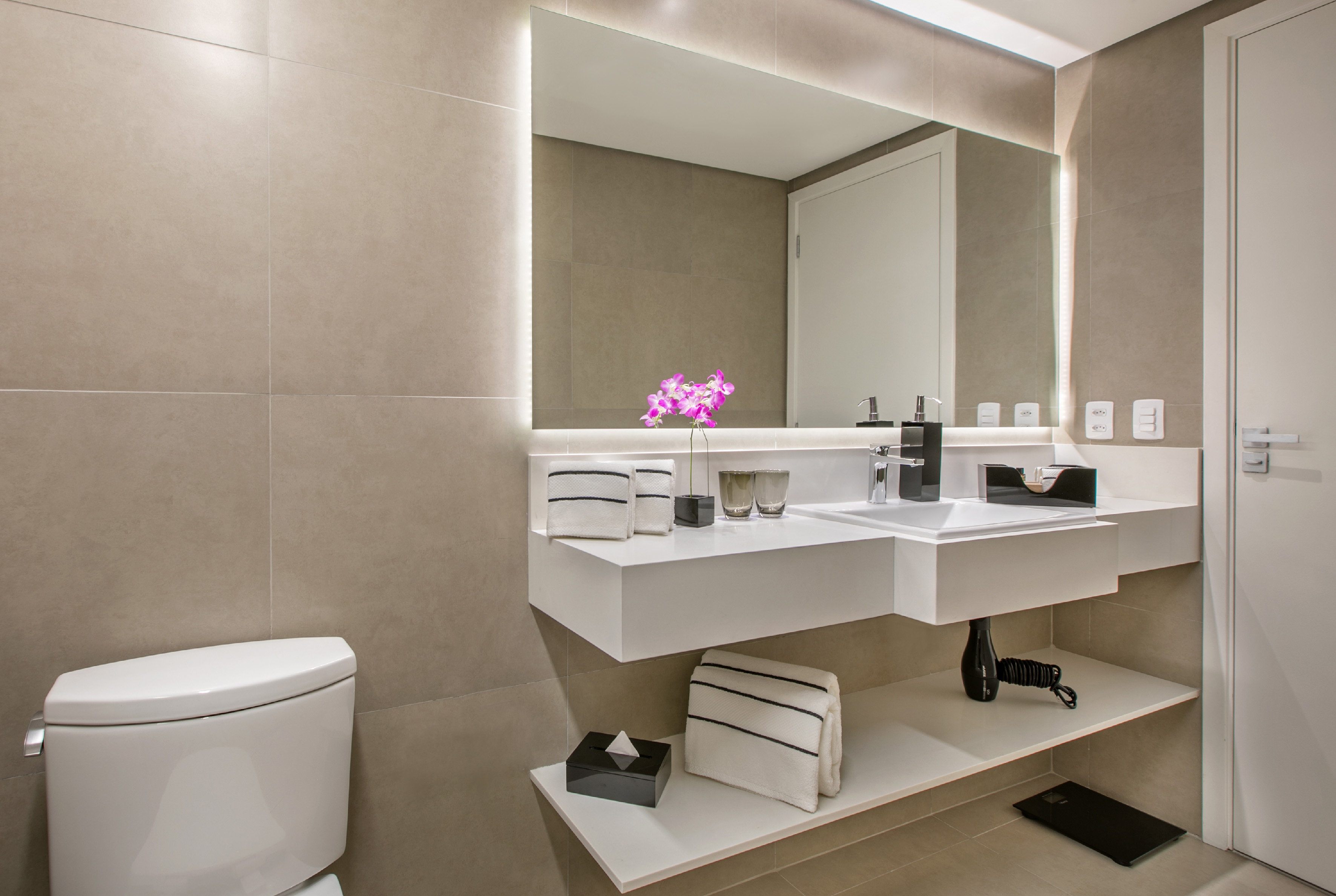 Minimalist hotel bathroom with white floating vanity, large lighted mirror, and toilet.