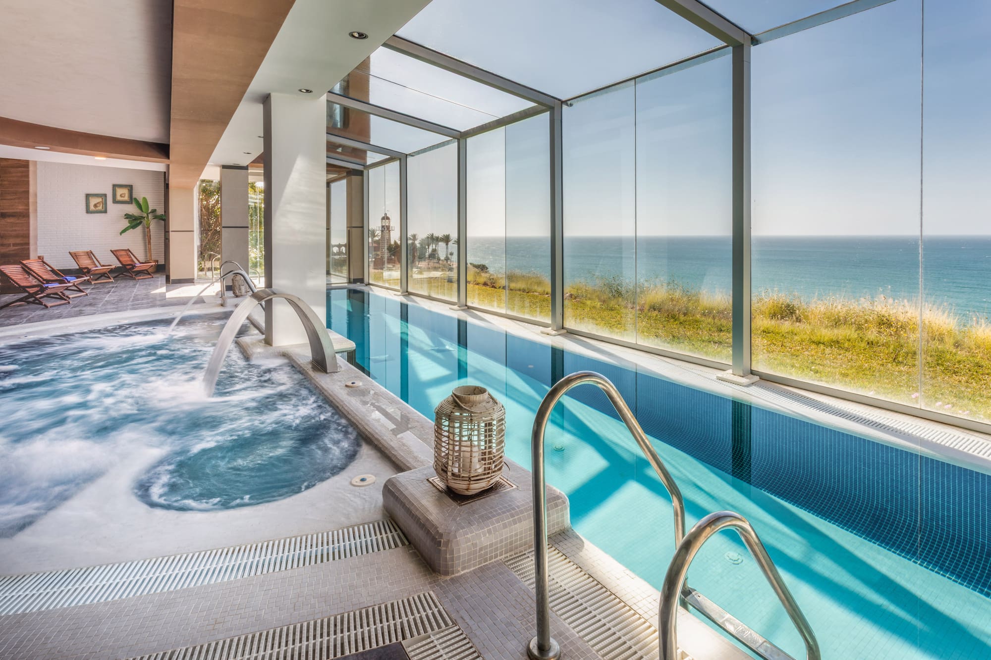 Coastal indoor swimming pool with sea view, jacuzzi, and relaxing lounge area.