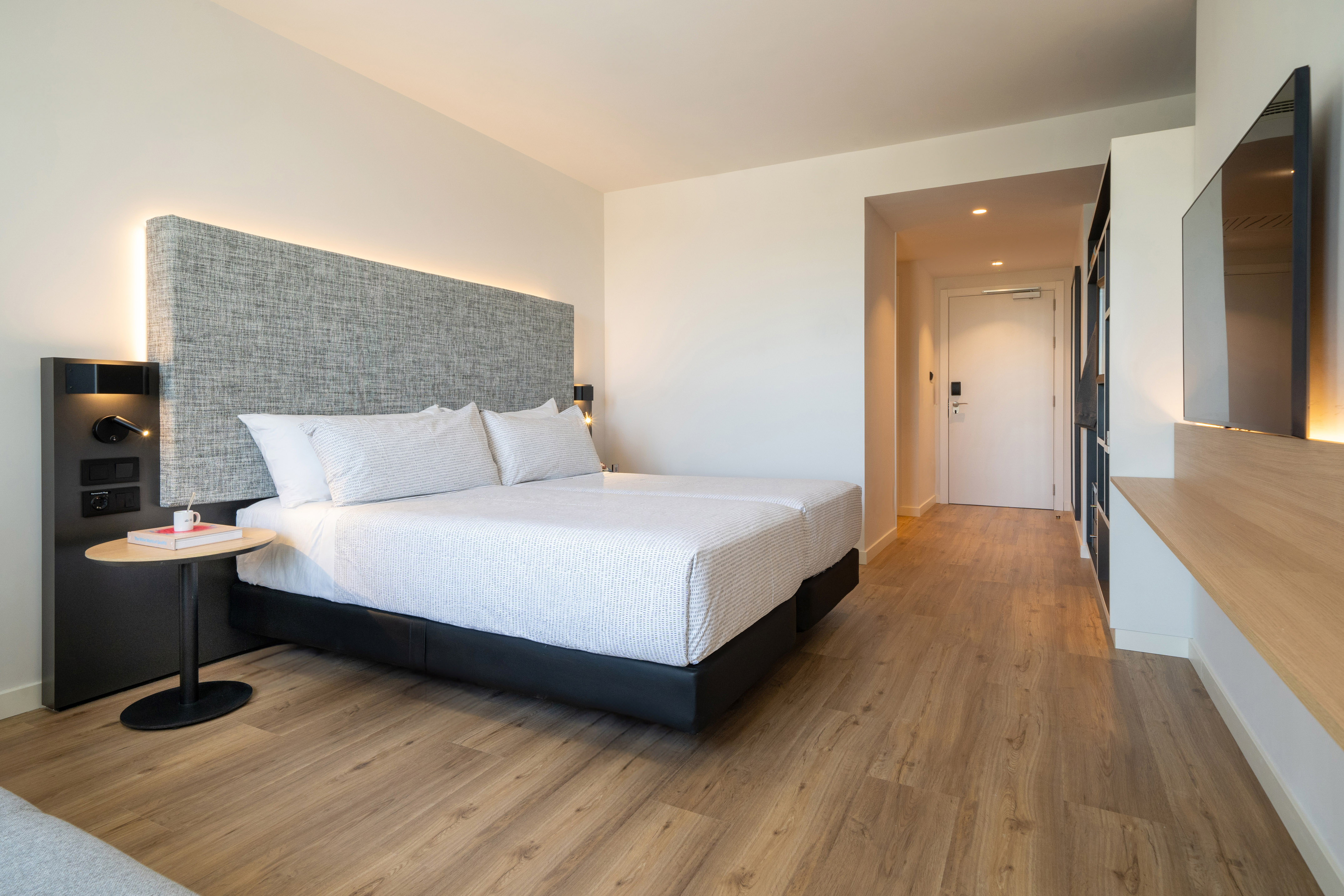 Modern hotel room with backlit grey headboard, twin beds, and wooden floor.