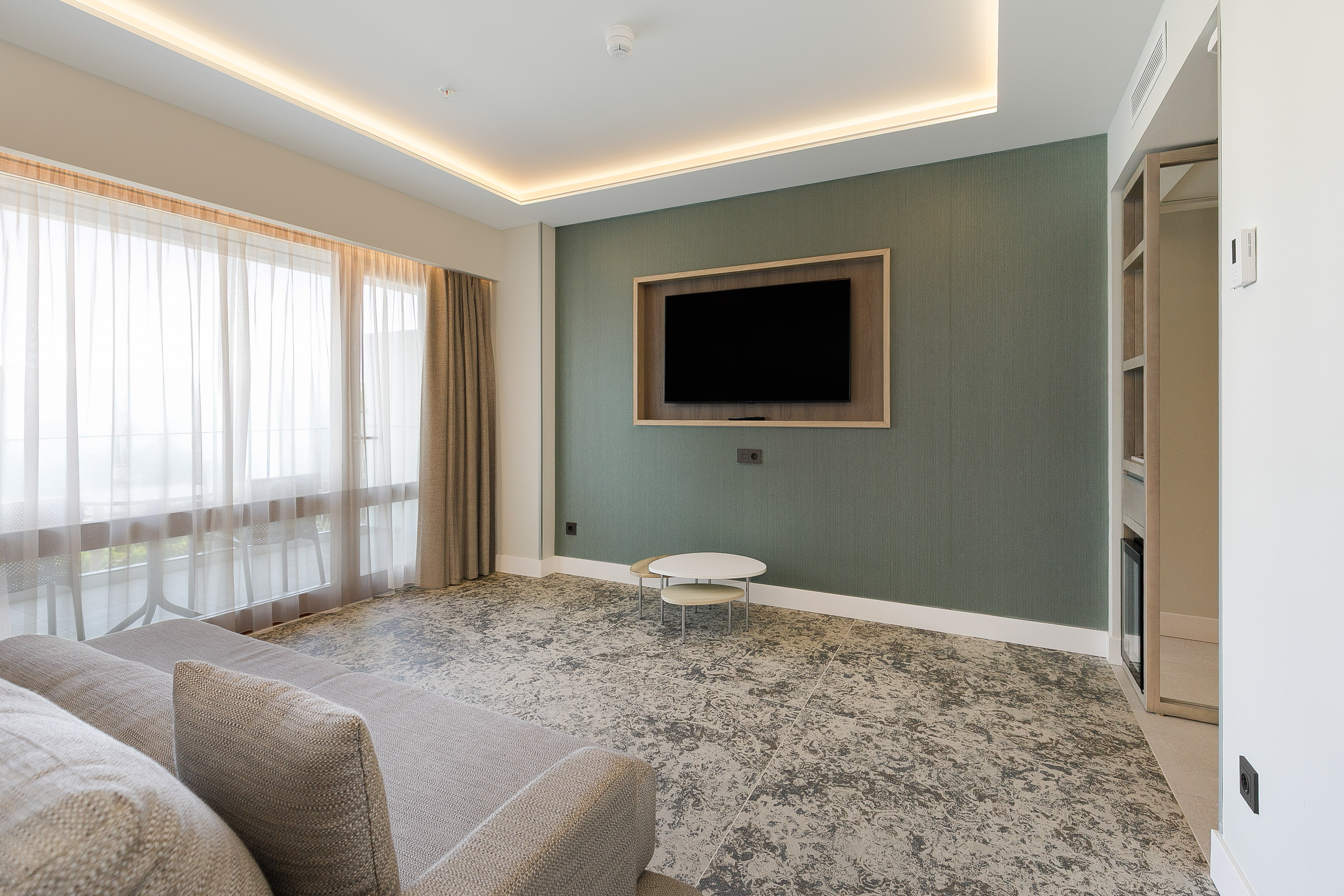 Modern hotel living room with gray sofa, large window, TV on green wall.