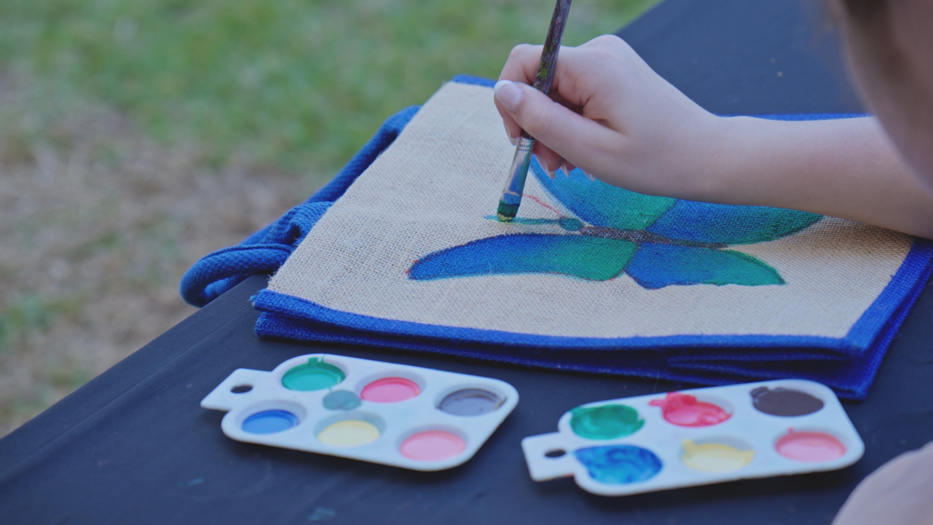Hand painting a blue butterfly on burlap fabric, with paint palettes and brush.