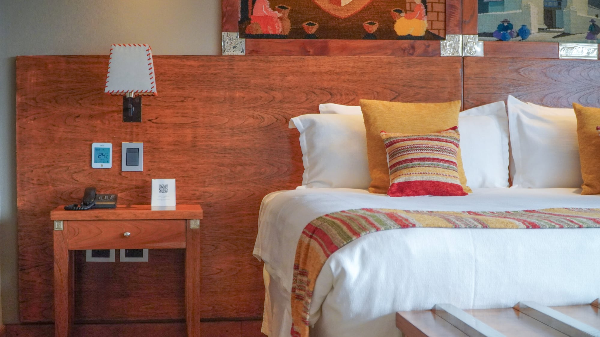 Hotel room with bed, wooden headboard, colorful pillows, nightstand, and tapestry.