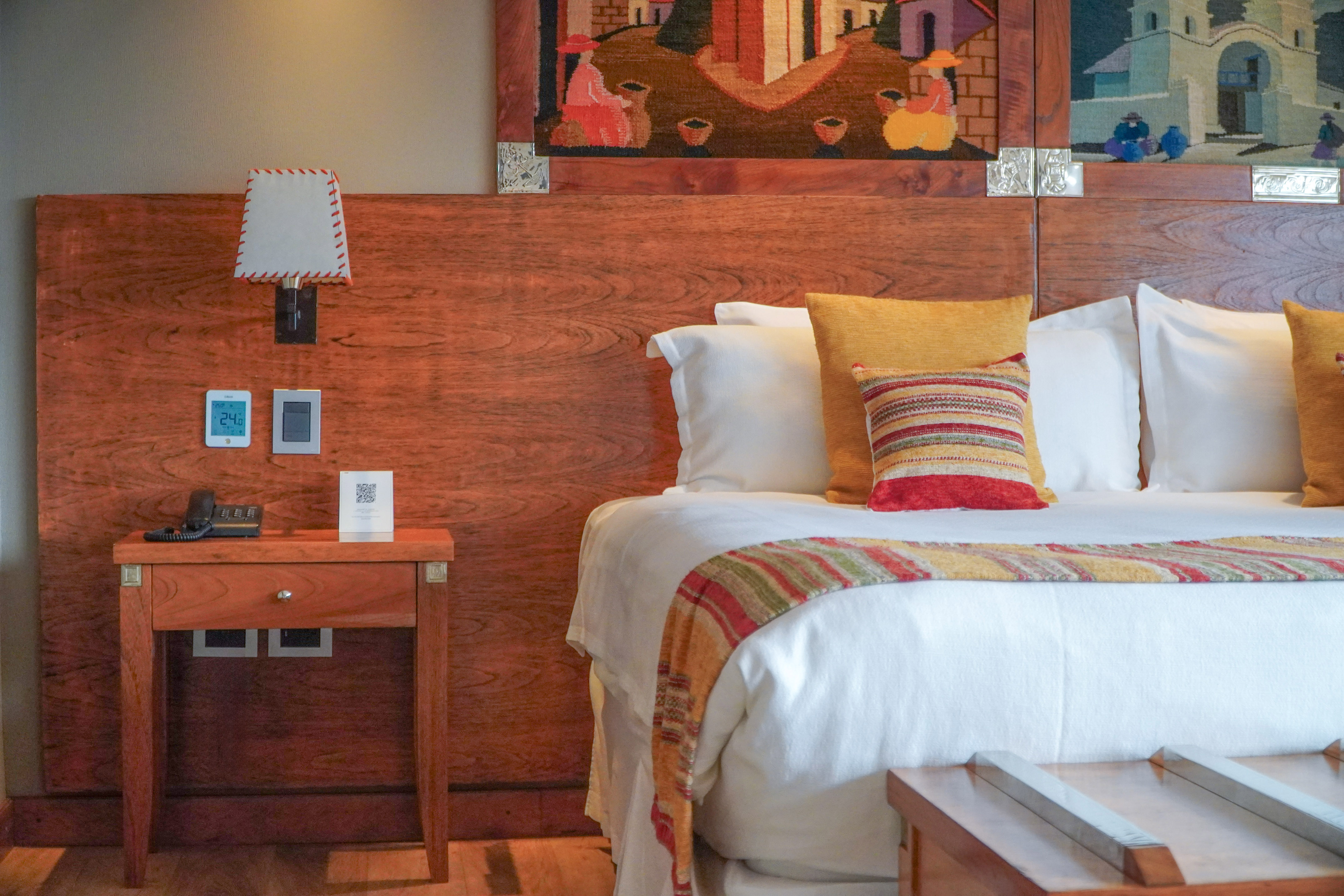 Hotel room with bed, wooden headboard, colorful pillows, nightstand, and tapestry.