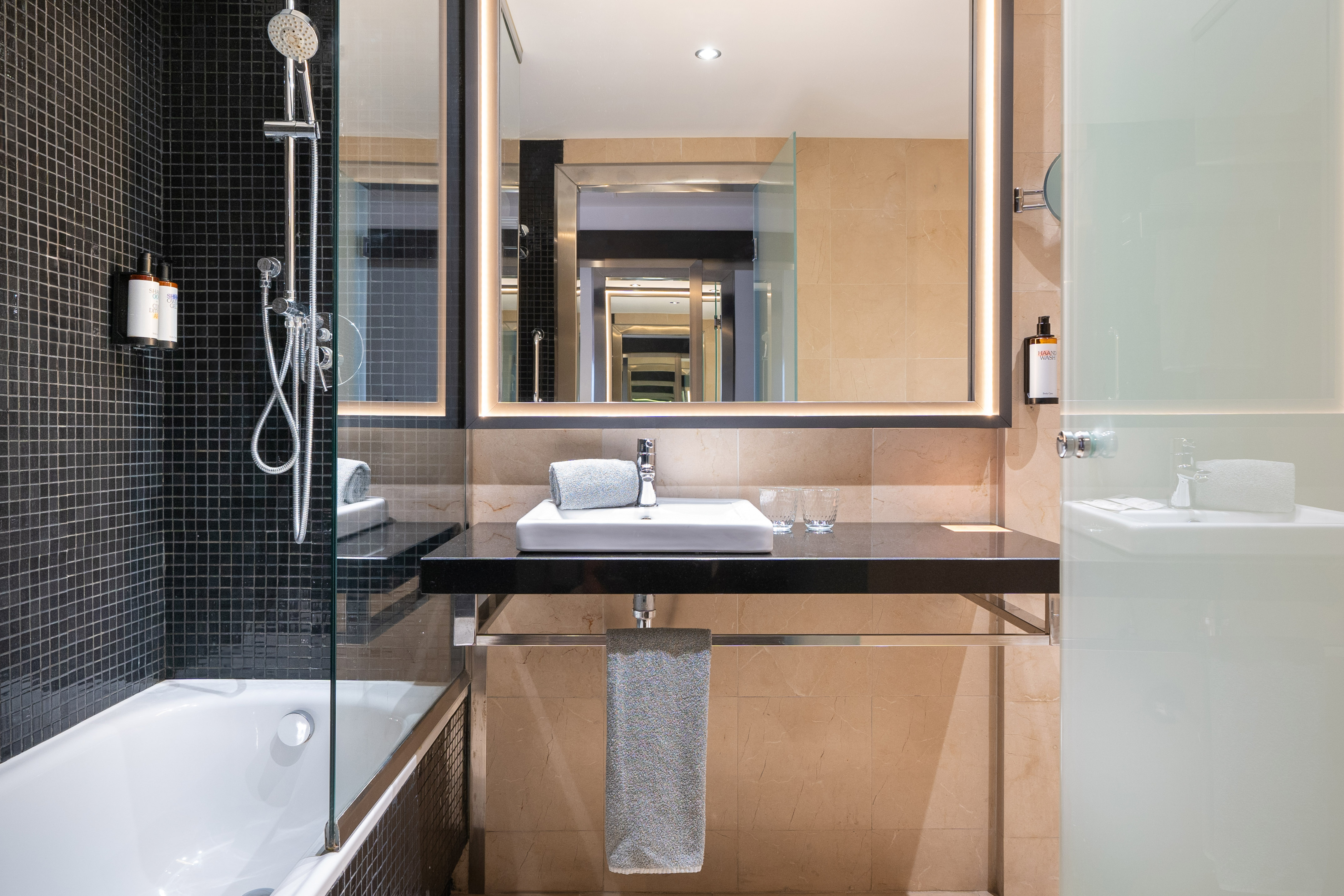 Luxury hotel bathroom with black tiled shower-tub, modern vanity, and illuminated mirror.
