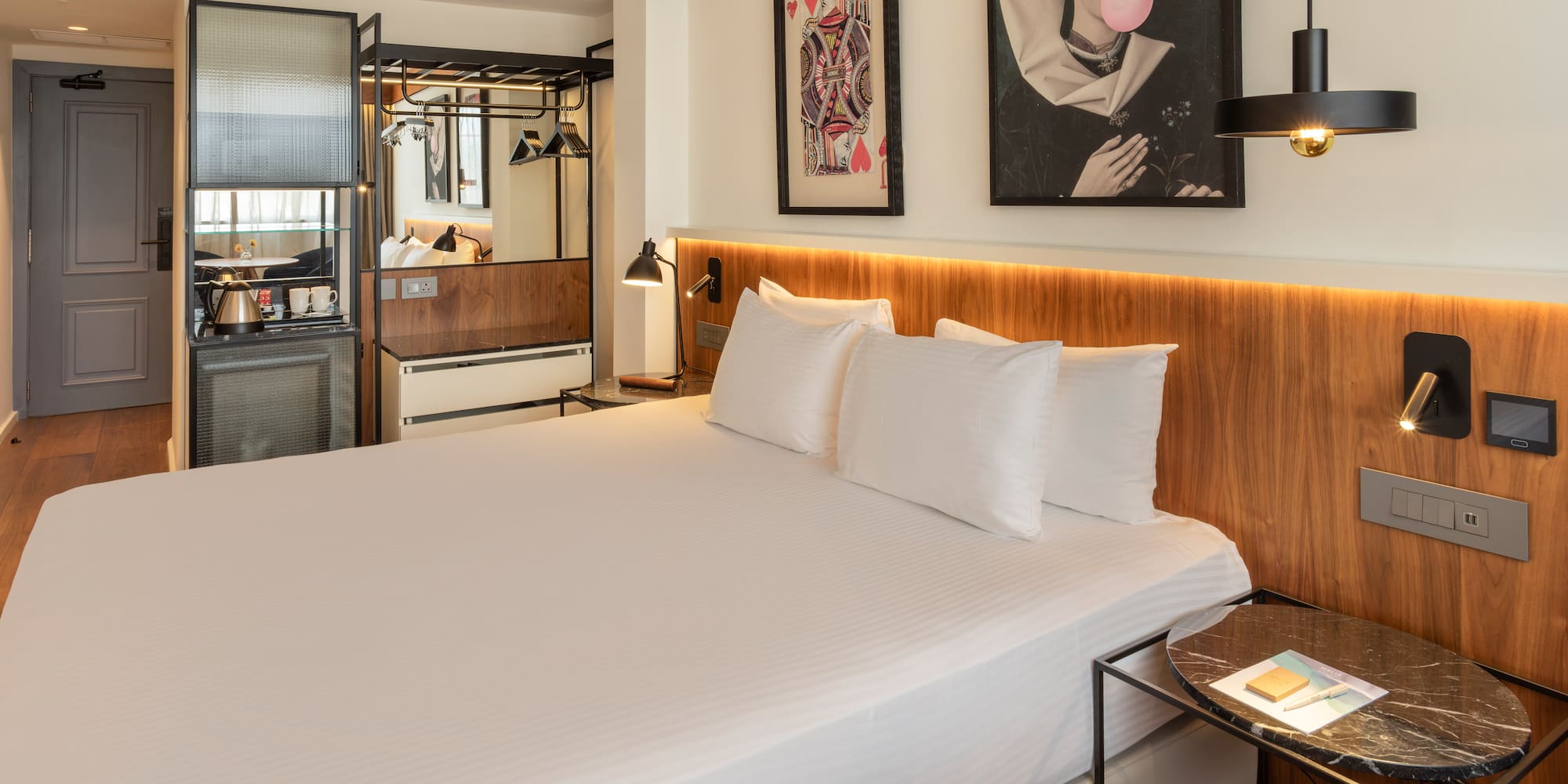 Modern hotel room with king bed, wooden headboard, minibar, and eclectic art.