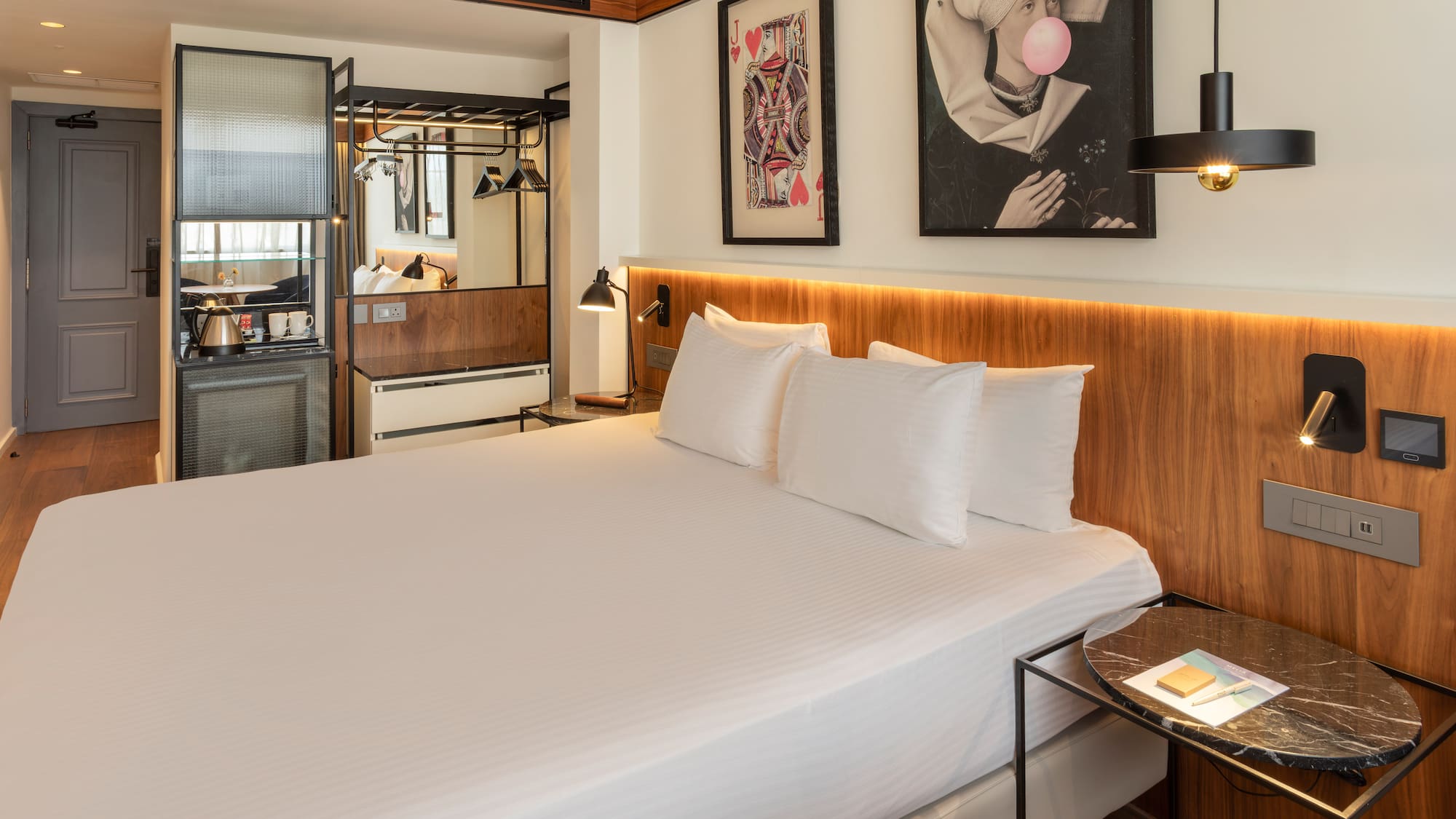 Modern hotel room with king bed, wooden headboard, minibar, and eclectic art.