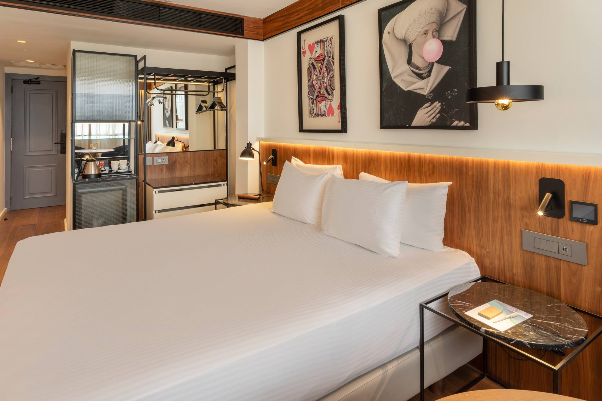 Modern hotel room with king bed, wooden headboard, minibar, and eclectic art.