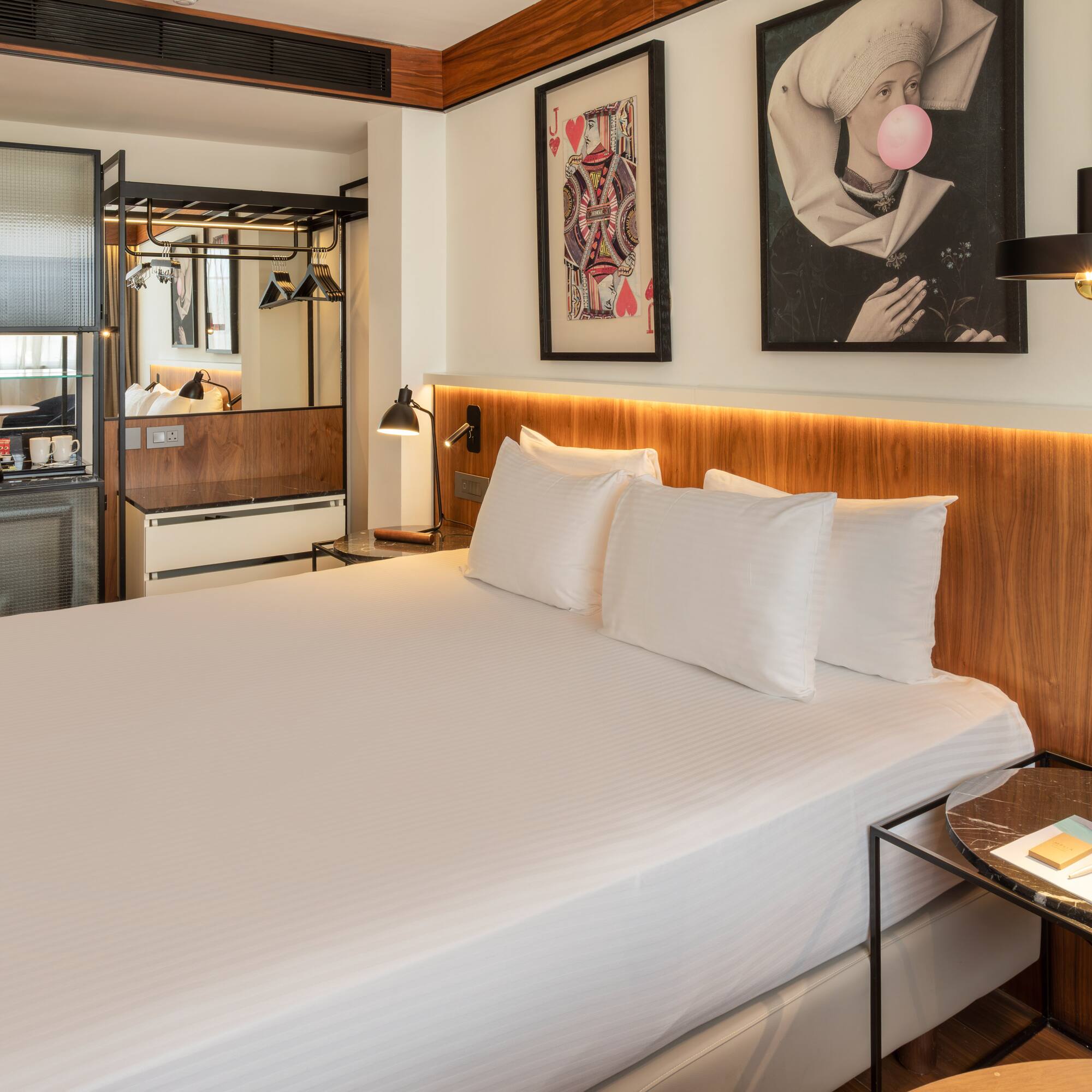 Modern hotel room with king bed, wooden headboard, minibar, and eclectic art.