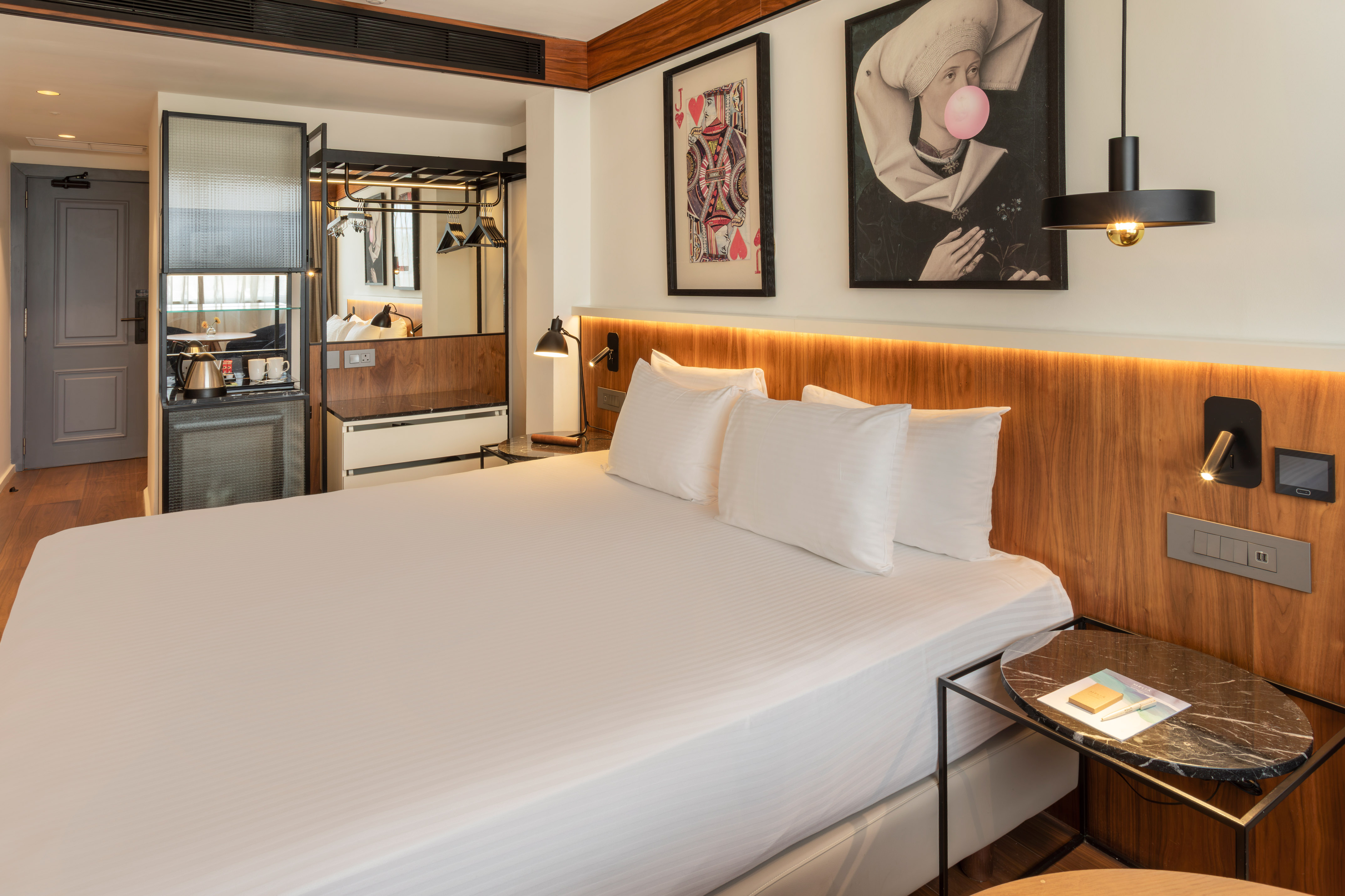Modern hotel room with king bed, wooden headboard, minibar, and eclectic art.