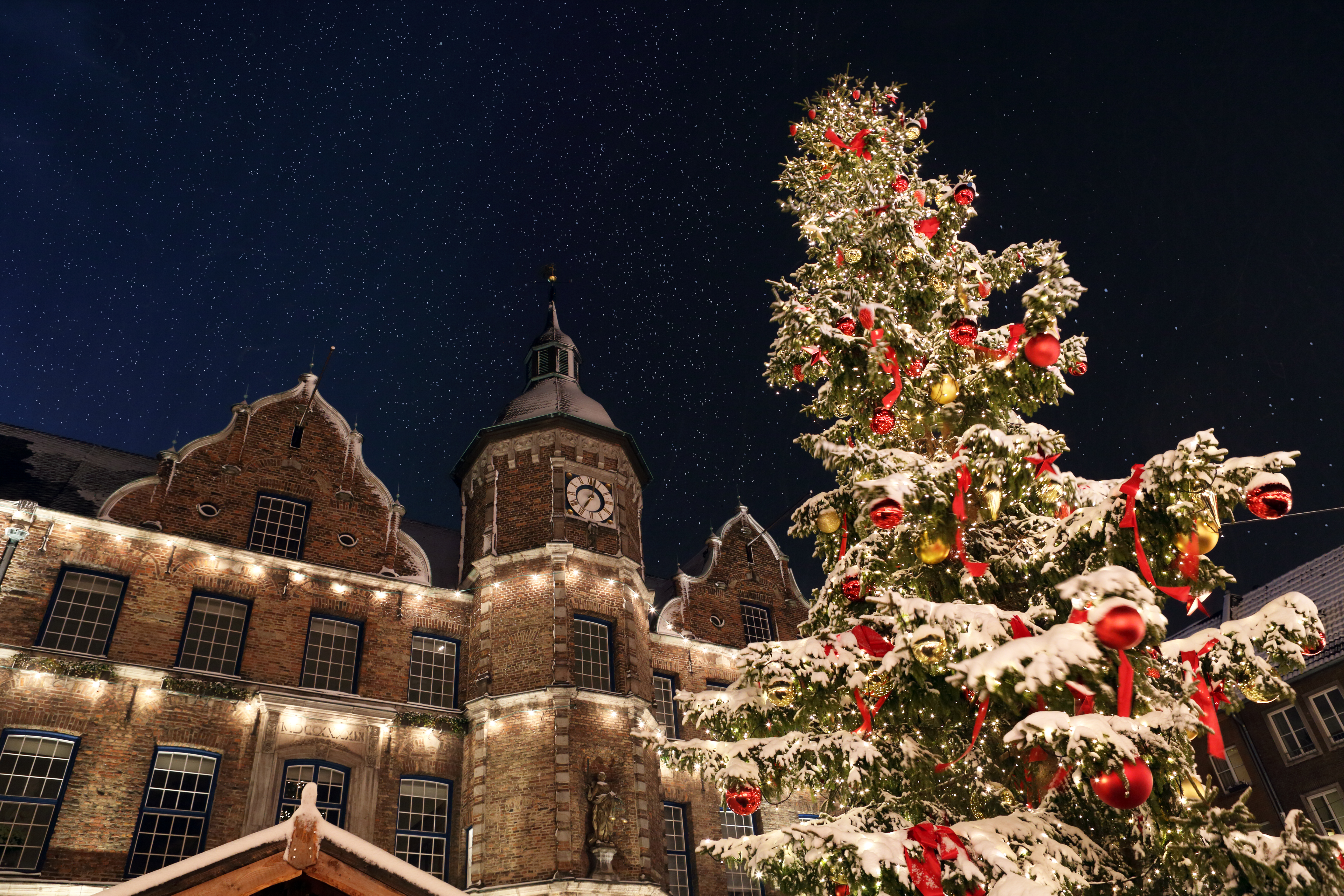 Snowy Christmas tree with ornaments, historic clock tower, and starry night sky.