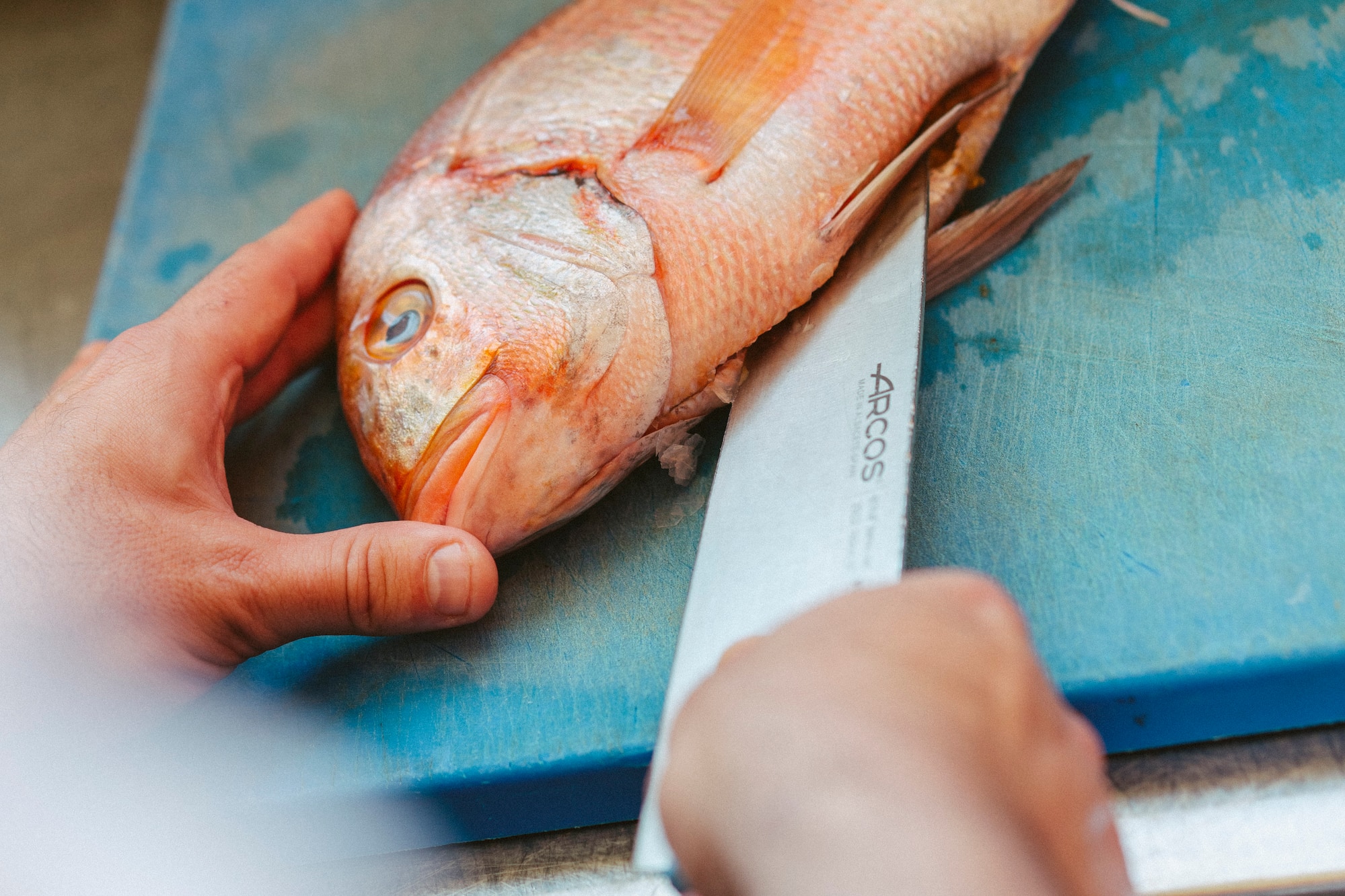 Chef preparing fresh red snapper with knife on blue cutting board.