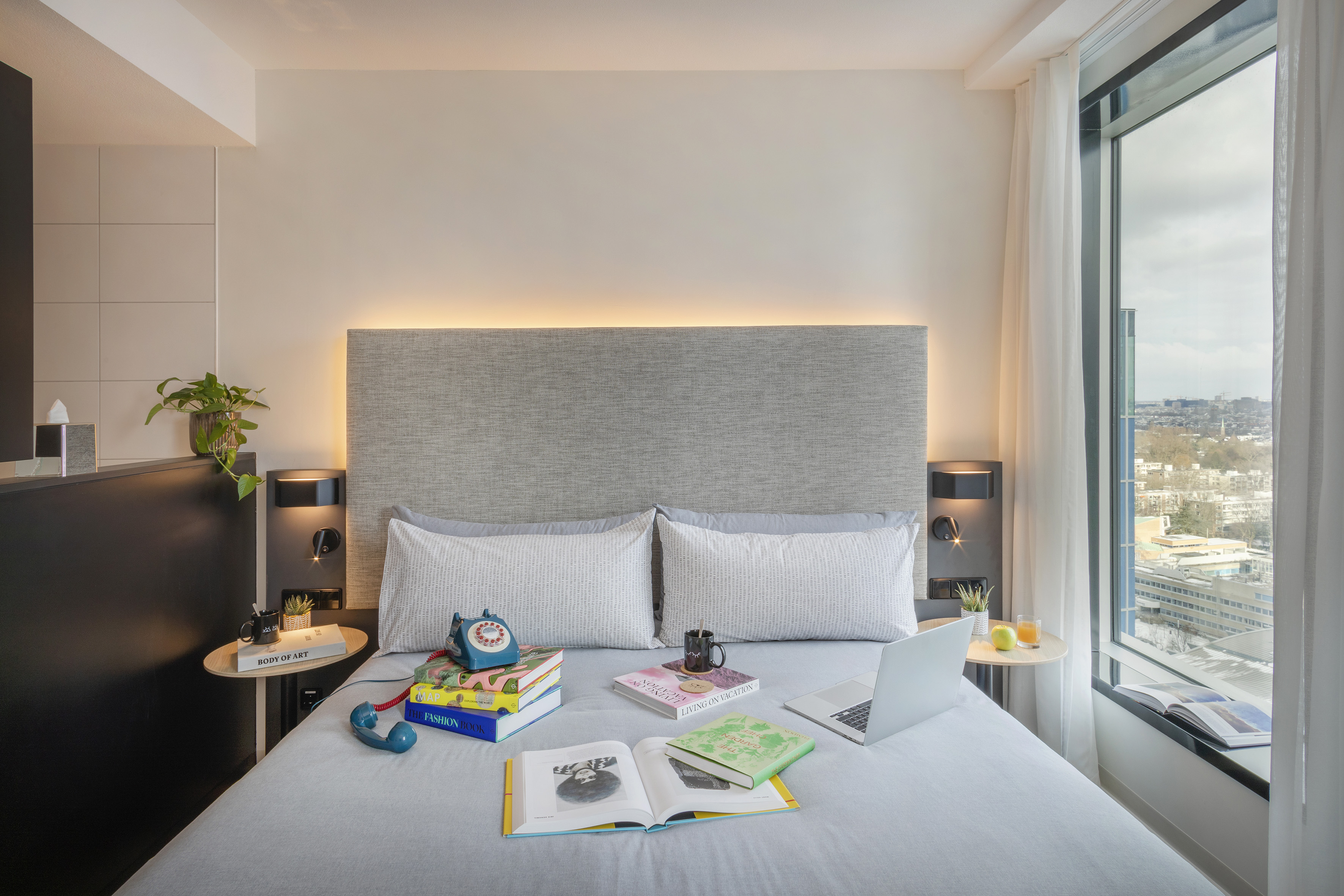 Contemporary hotel room: bed, backlit headboard, laptop, books, city view.