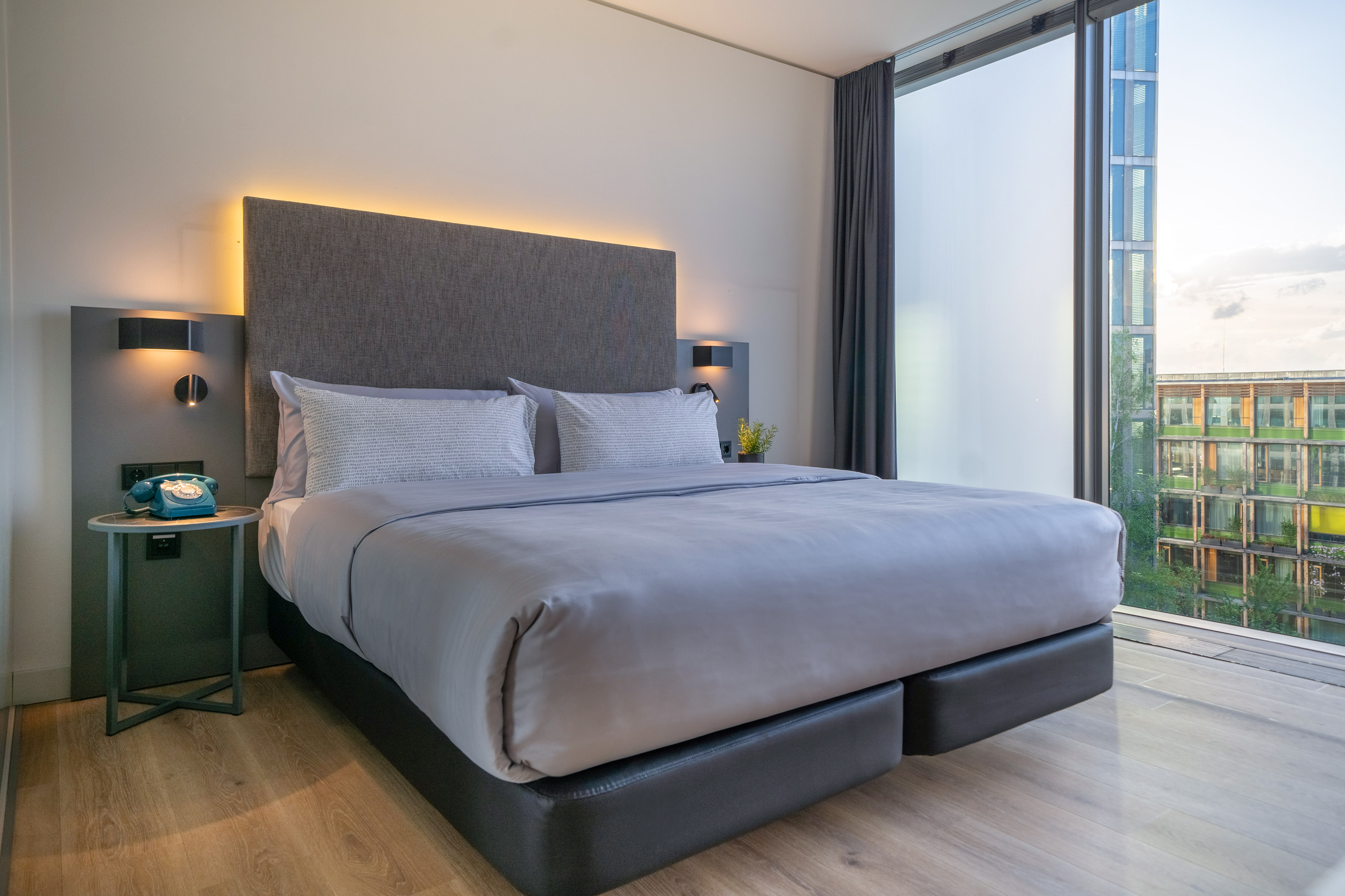 Modern hotel room with king bed, grey headboard, backlit, city view window.