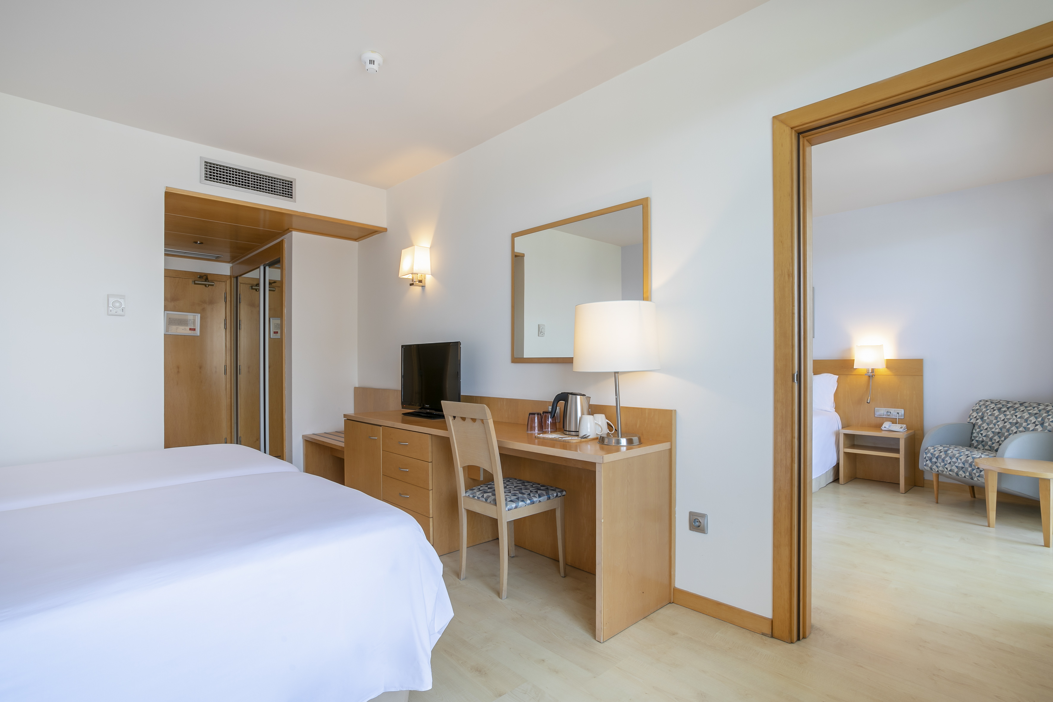 Modern hotel room with twin beds, desk, TV, and an adjacent lounge area.