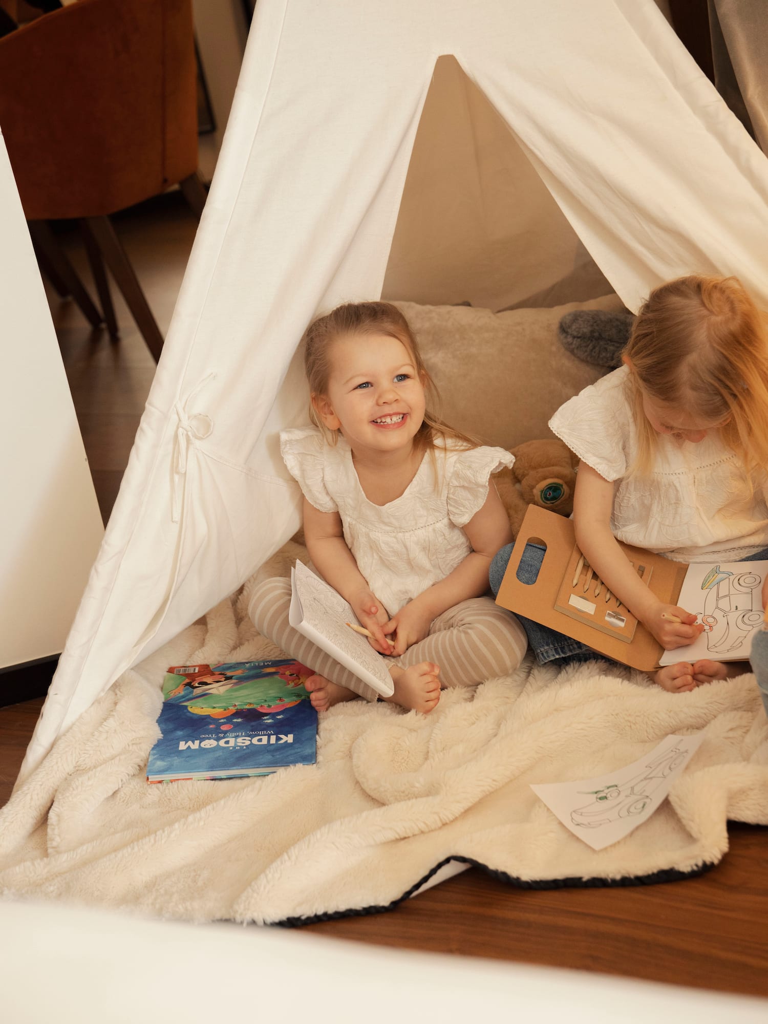 Toddler girls playing, coloring in a cozy white teepee tent with books.