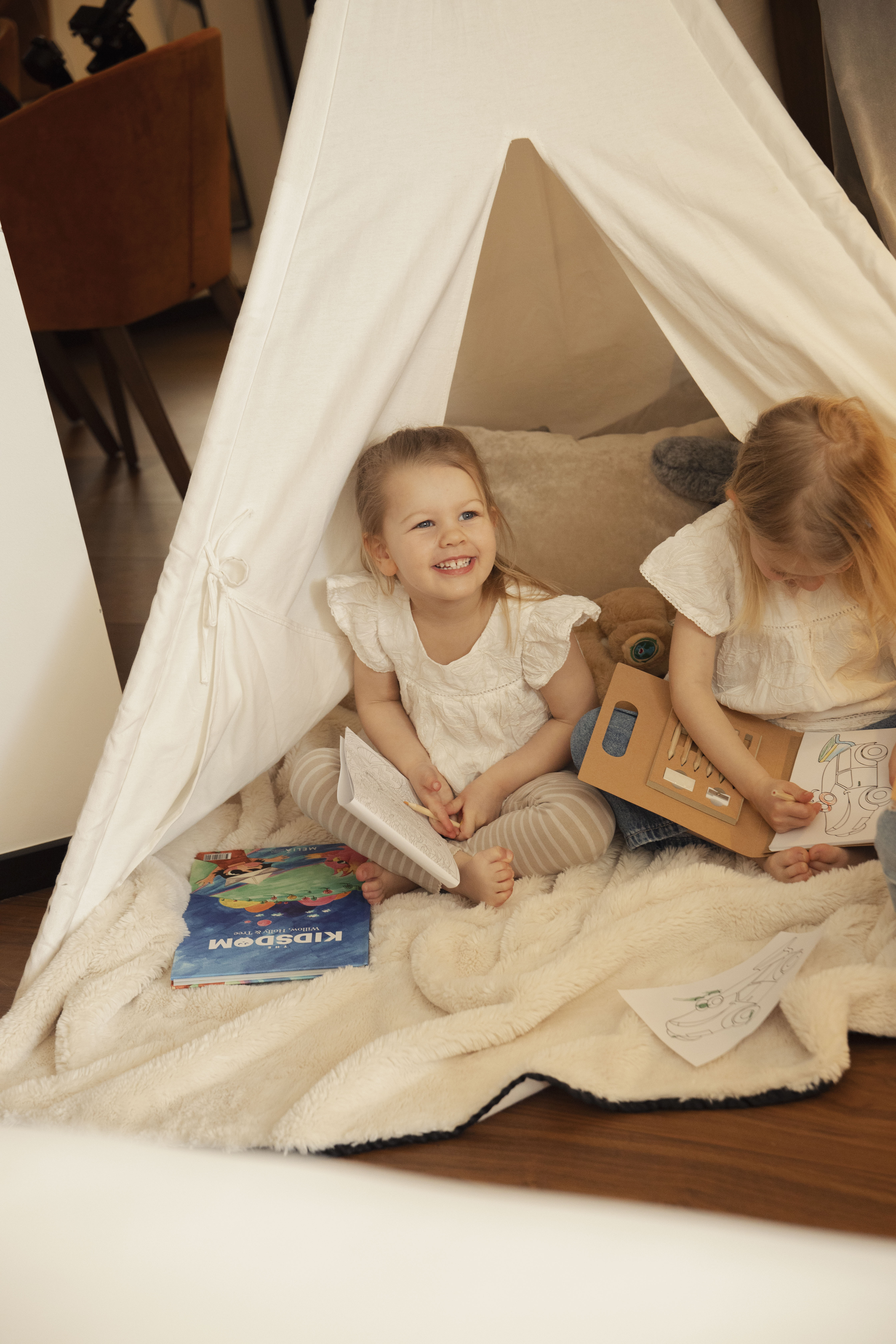 Toddler girls playing, coloring in a cozy white teepee tent with books.