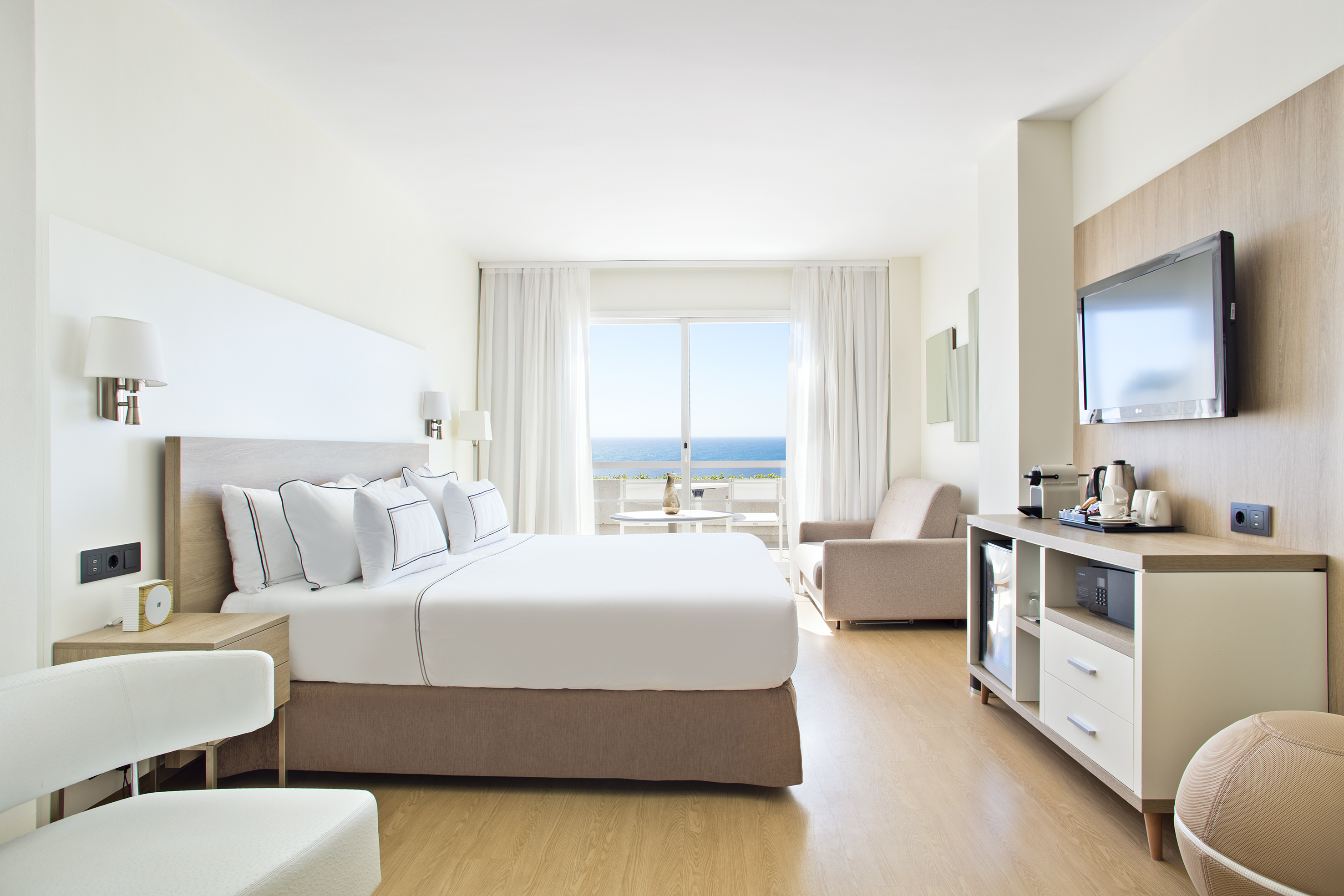 Modern hotel room with ocean view, king bed, TV, and mini-bar.