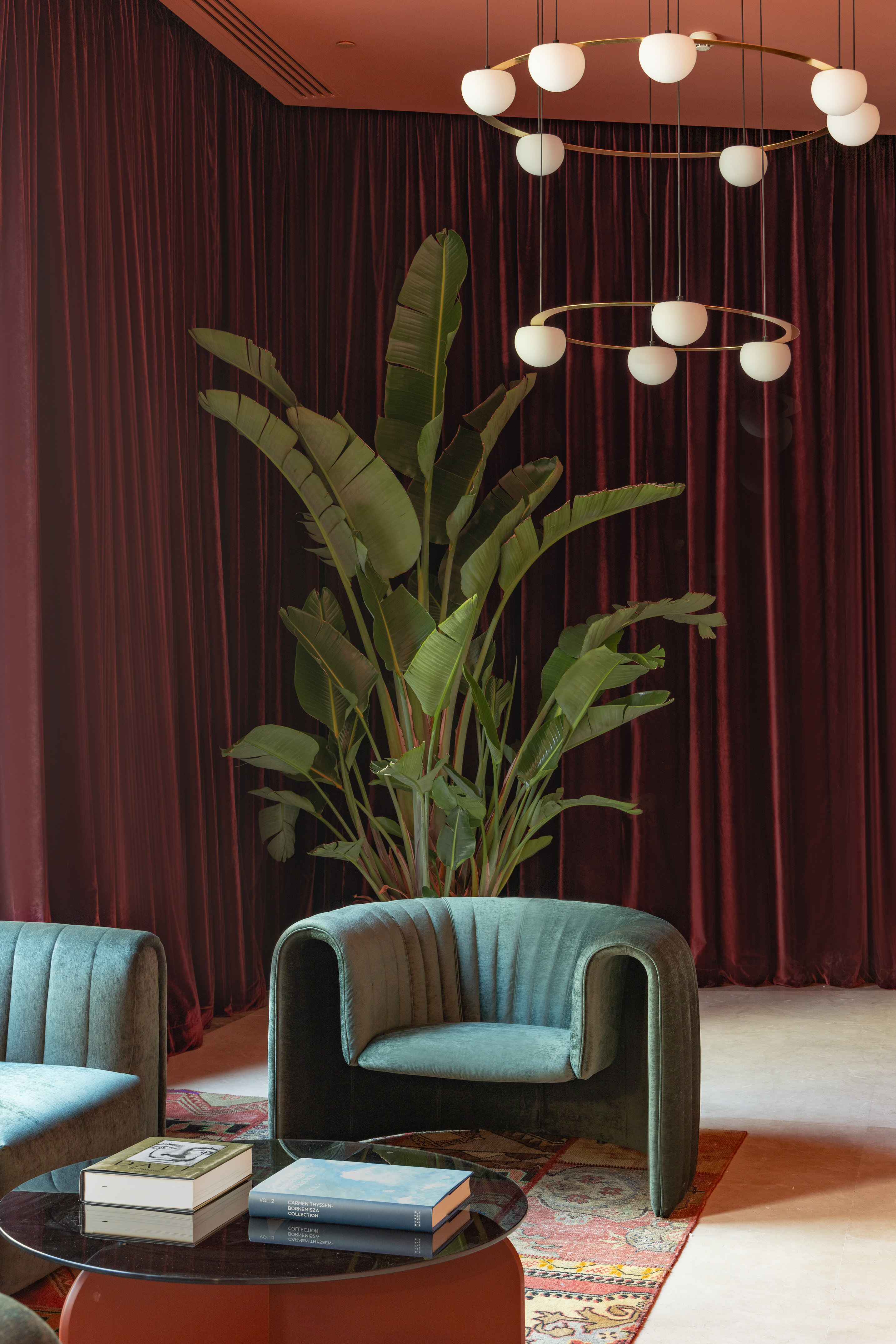 Modern lounge: green velvet chairs, large plant, red curtains, globe chandelier.