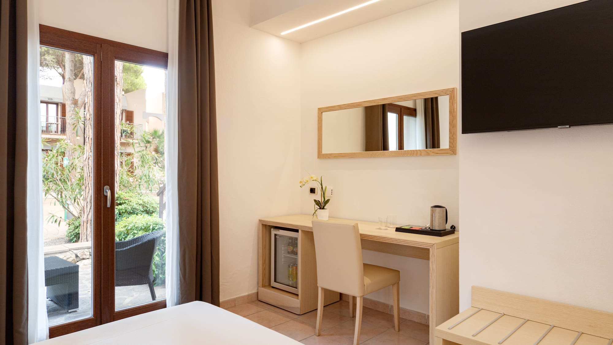 Modern hotel bedroom with desk, mini-fridge, wall TV, and patio view.