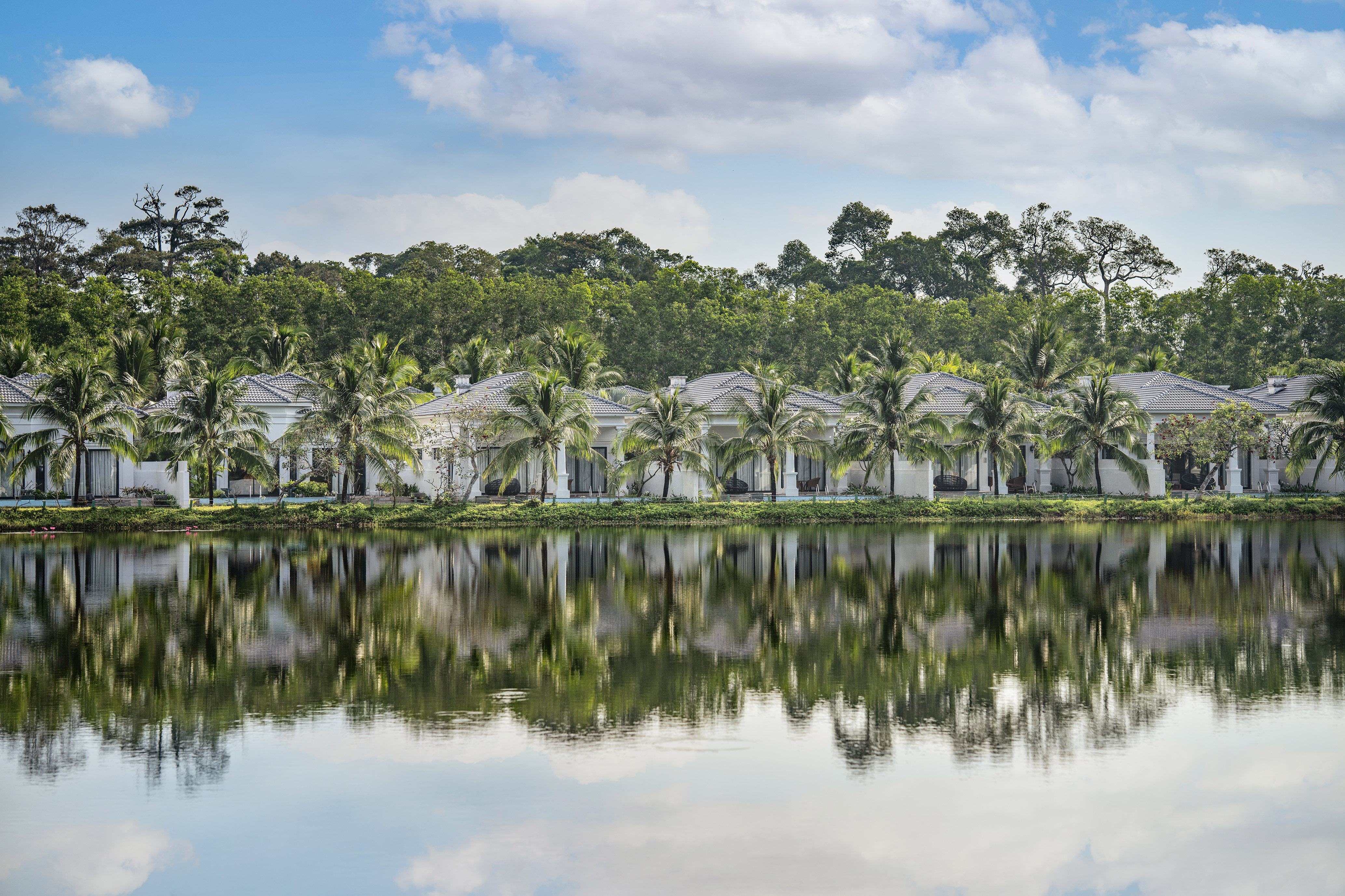 Tropical resort villas with palm trees reflecting in a serene lake.