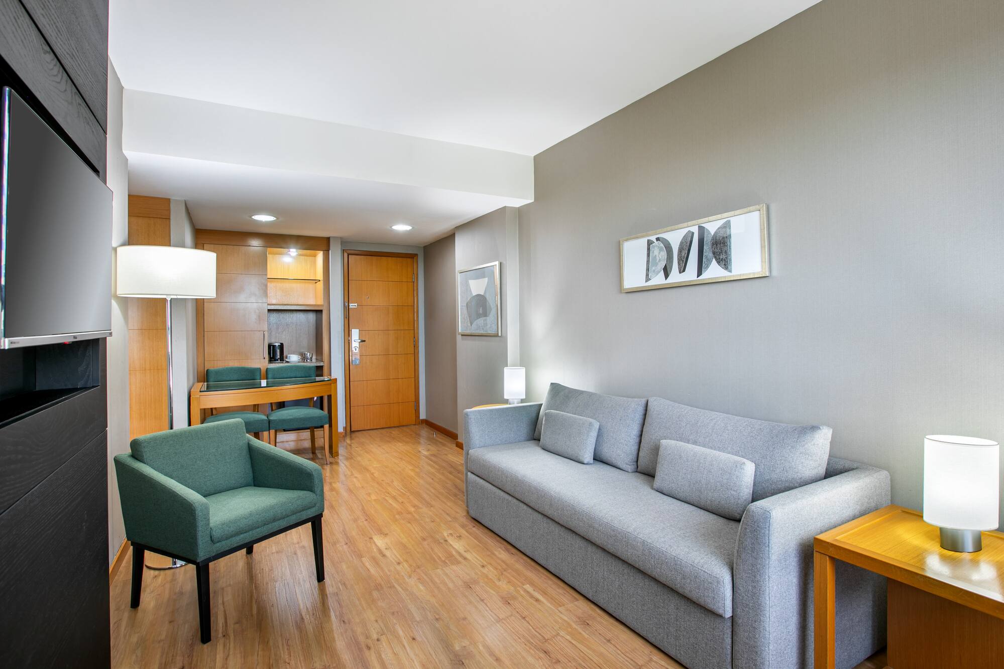 Hotel suite living area featuring grey sofa, green armchair, TV, and dining nook.