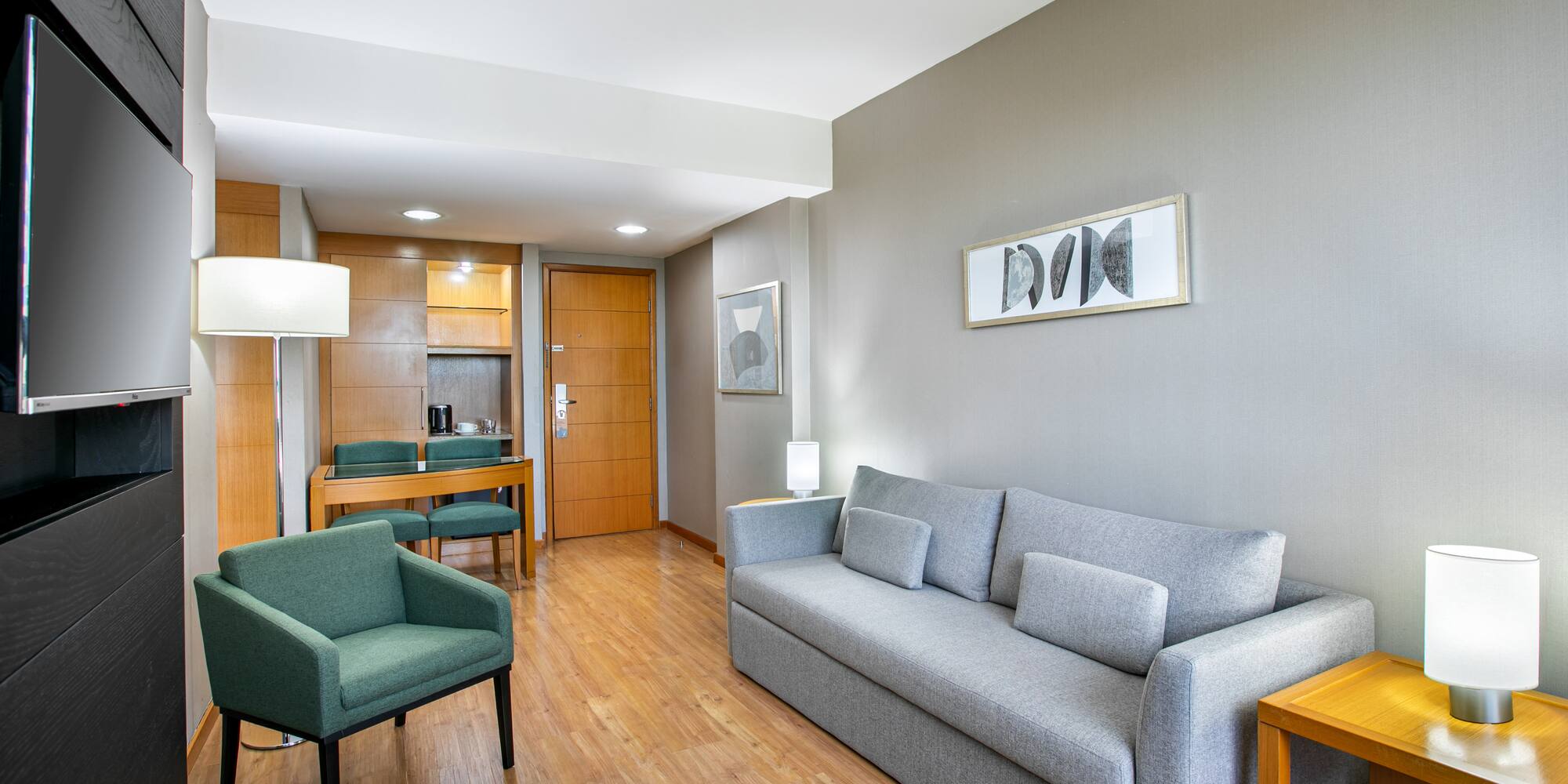 Hotel suite living area featuring grey sofa, green armchair, TV, and dining nook.
