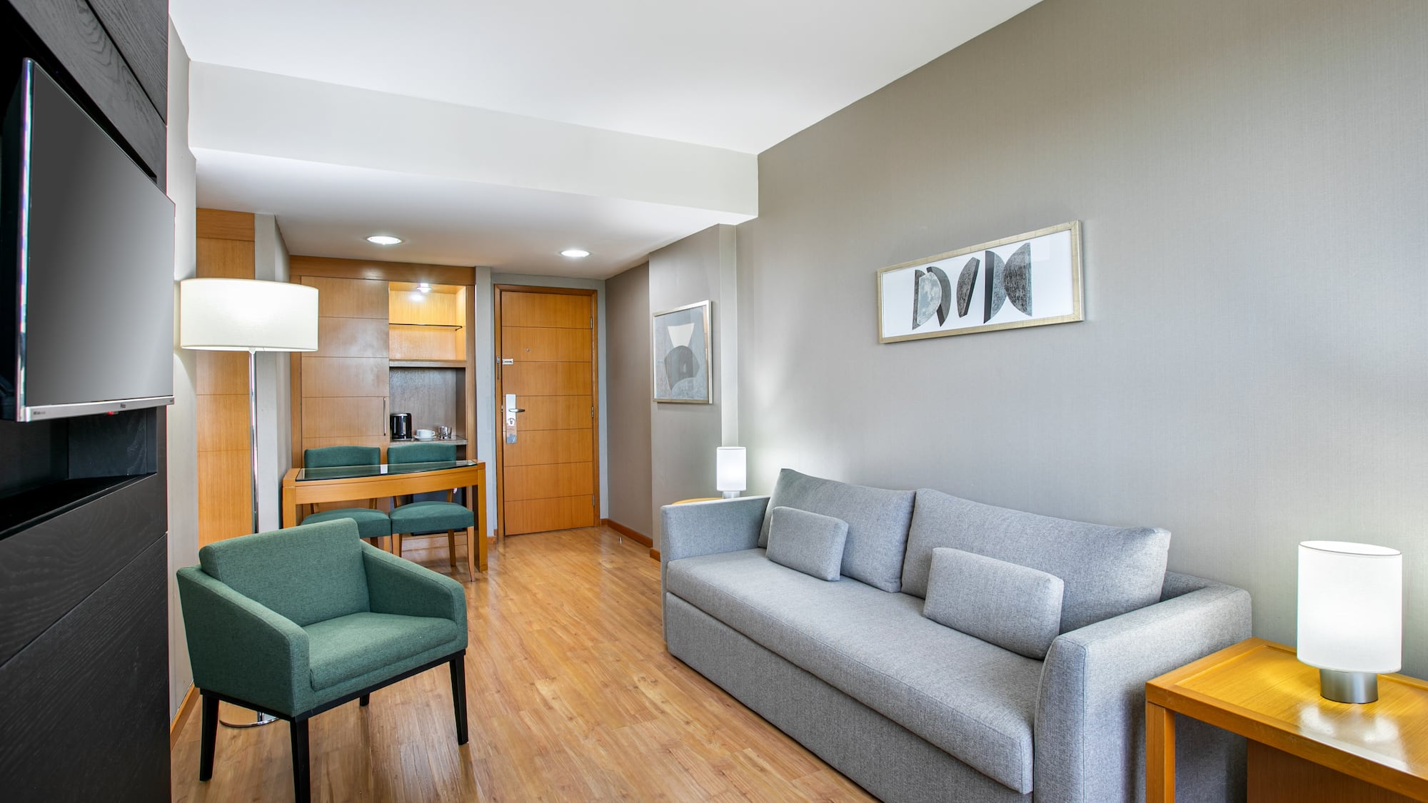 Hotel suite living area featuring grey sofa, green armchair, TV, and dining nook.