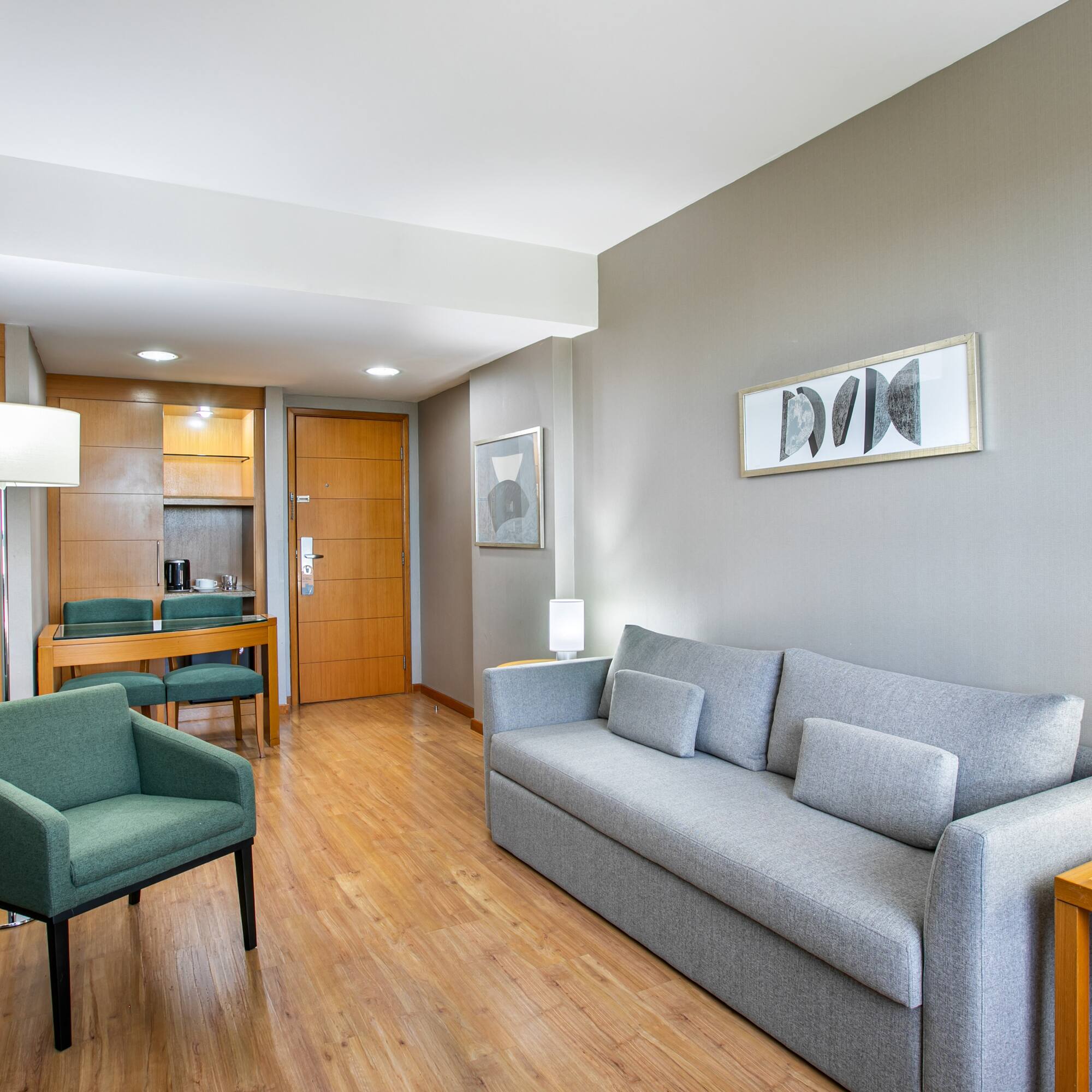Hotel suite living area featuring grey sofa, green armchair, TV, and dining nook.